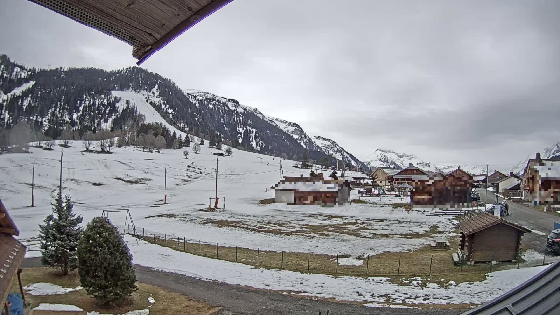 A snowy mountain village is seen under a cloudy sky, with patches of snow covering the ground and some bare trees.