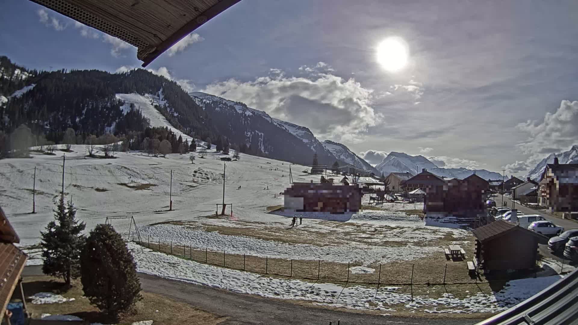 A sunny day with partly cloudy skies overlooks a snow-dusted village nestled in a valley between mountains, with a few cars parked and a small playground visible.