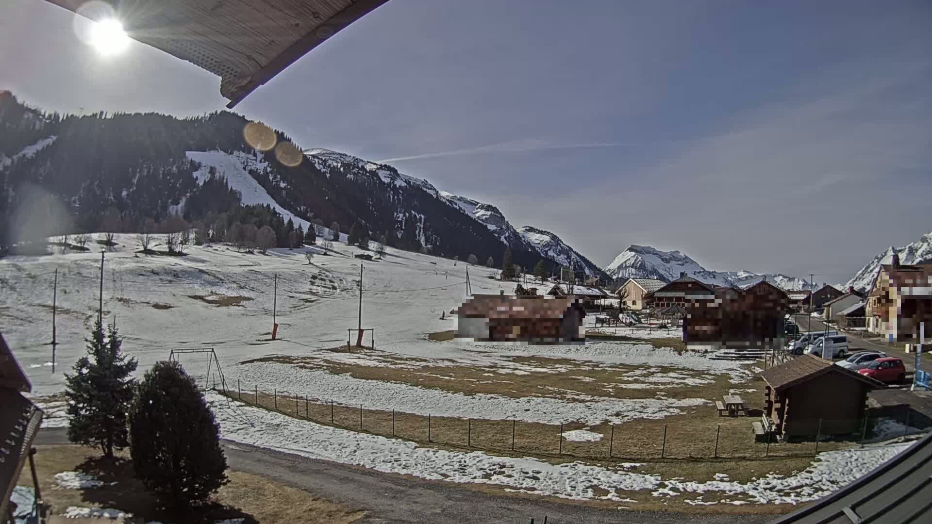 A sunny day in a snow-dusted mountain village, with patchy snow covering the ground and mountains in the background.