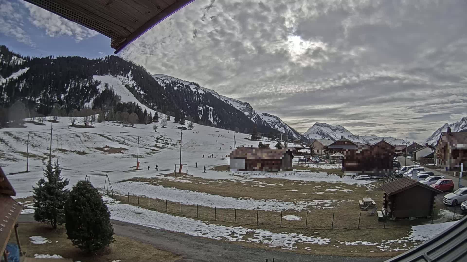 A partly cloudy day in a snow-covered mountainous region shows a small village with a few cars parked near some buildings, and a ski slope with several people skiing.
