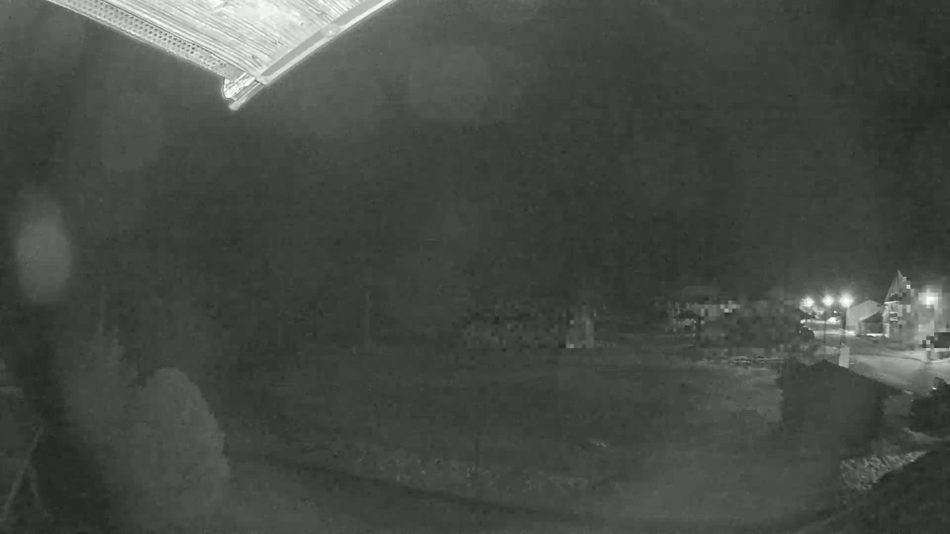 A nighttime, low-light outdoor scene shows a dark yard with dimly lit buildings and streetlights in the distance.