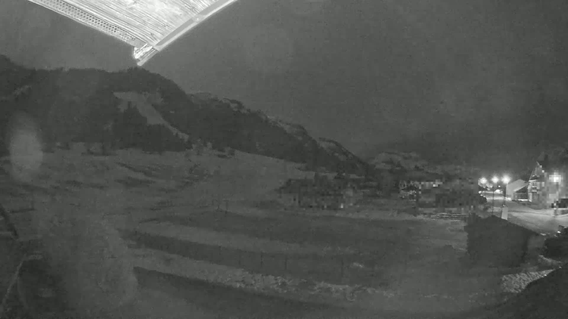 A nighttime, snow-covered landscape shows a small village nestled at the base of a dark mountain under a cloudy sky.