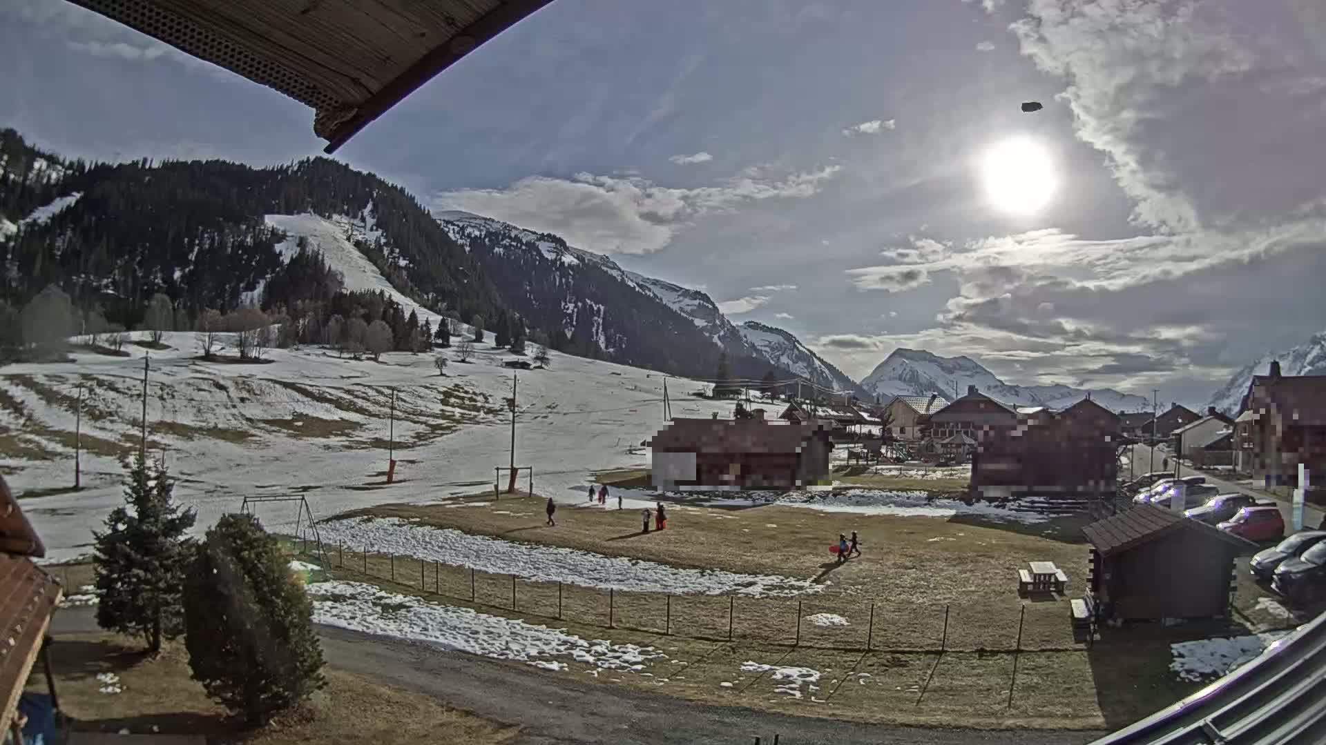 A sunny day with partly cloudy skies shows a snow-dusted mountain village with children playing in a grassy area, a few buildings, and a partially snow-covered hillside in the background.