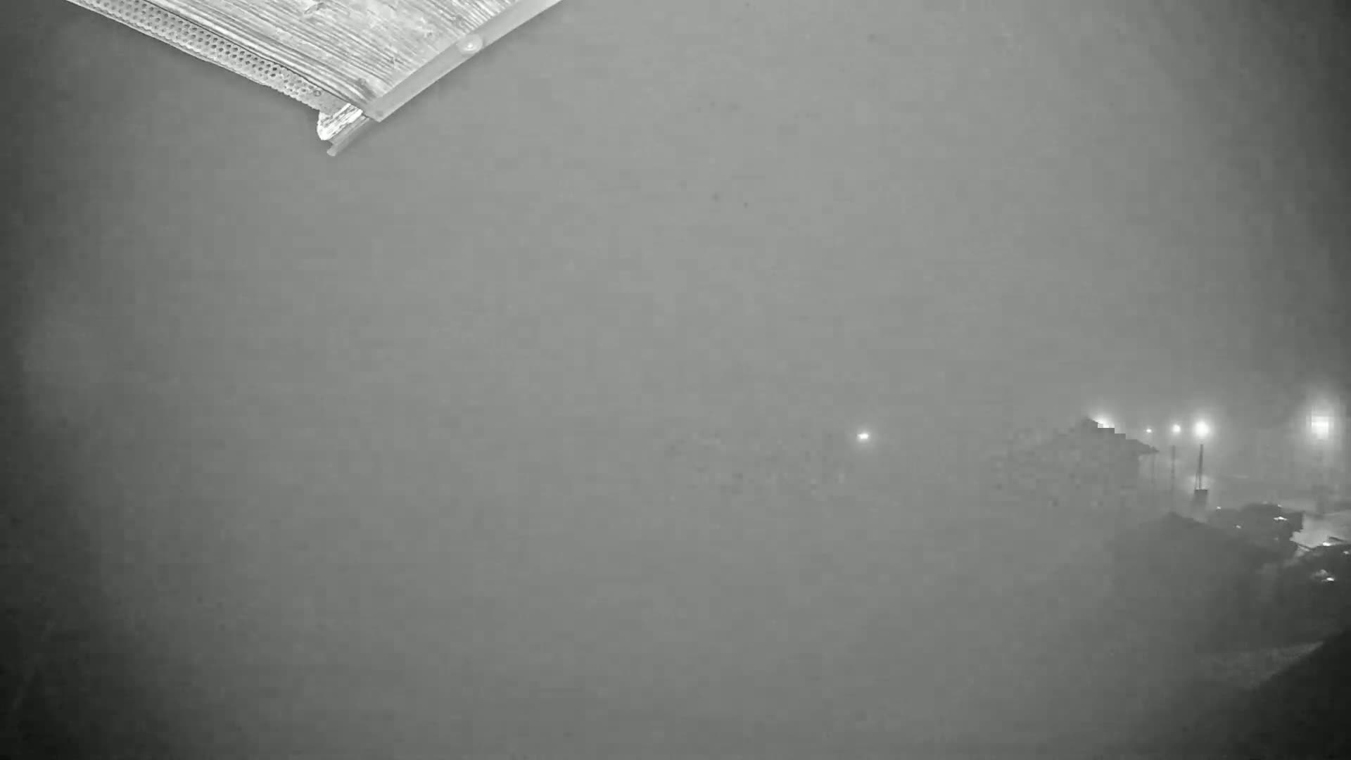 A nighttime, foggy view shows dimly lit buildings and streetlights in the distance, partially obscured by fog, from a perspective angled downward from a building's edge.