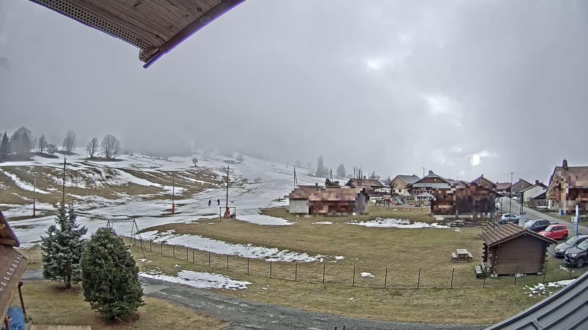 A mostly cloudy day reveals a small village nestled in a snow-dusted valley, with a few people visible on a gently sloping hillside.