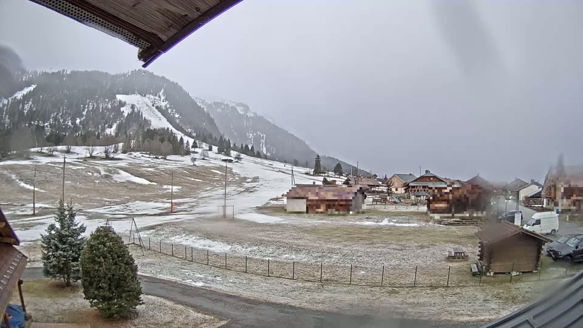 A small village nestled in a snow-dusted valley, with a mountain range in the background under an overcast sky.