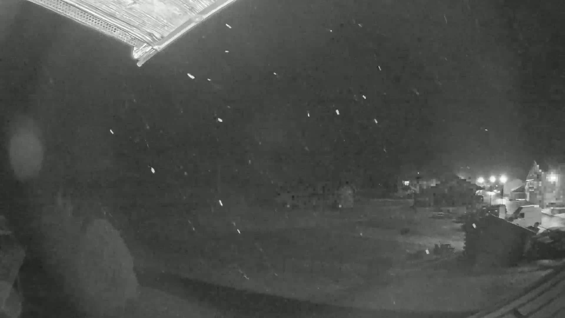 A nighttime, snowy scene is visible from a high angle, showing a mostly empty snow-covered area with some buildings and lights in the distance.