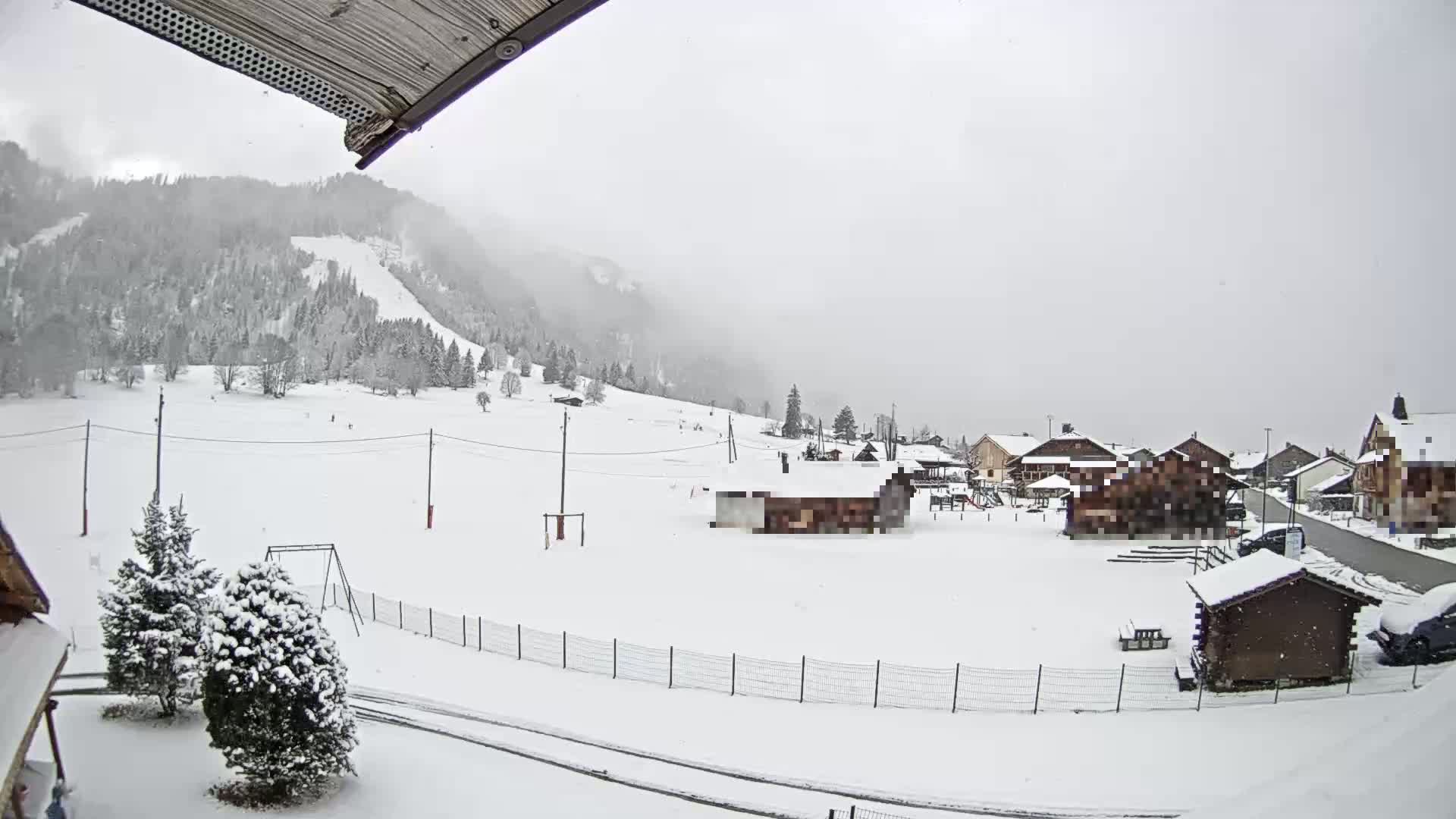 A snow-covered village nestled at the base of a snow-dusted mountain range under a gray, overcast sky with light snowfall.