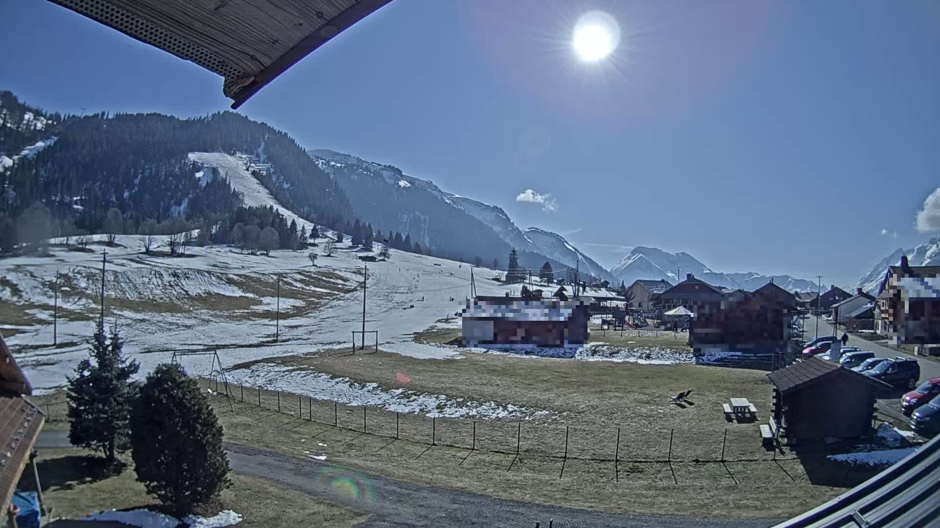 A sunny day in a snow-dusted mountain village with a few cars parked, some buildings, and a ski slope visible in the distance.