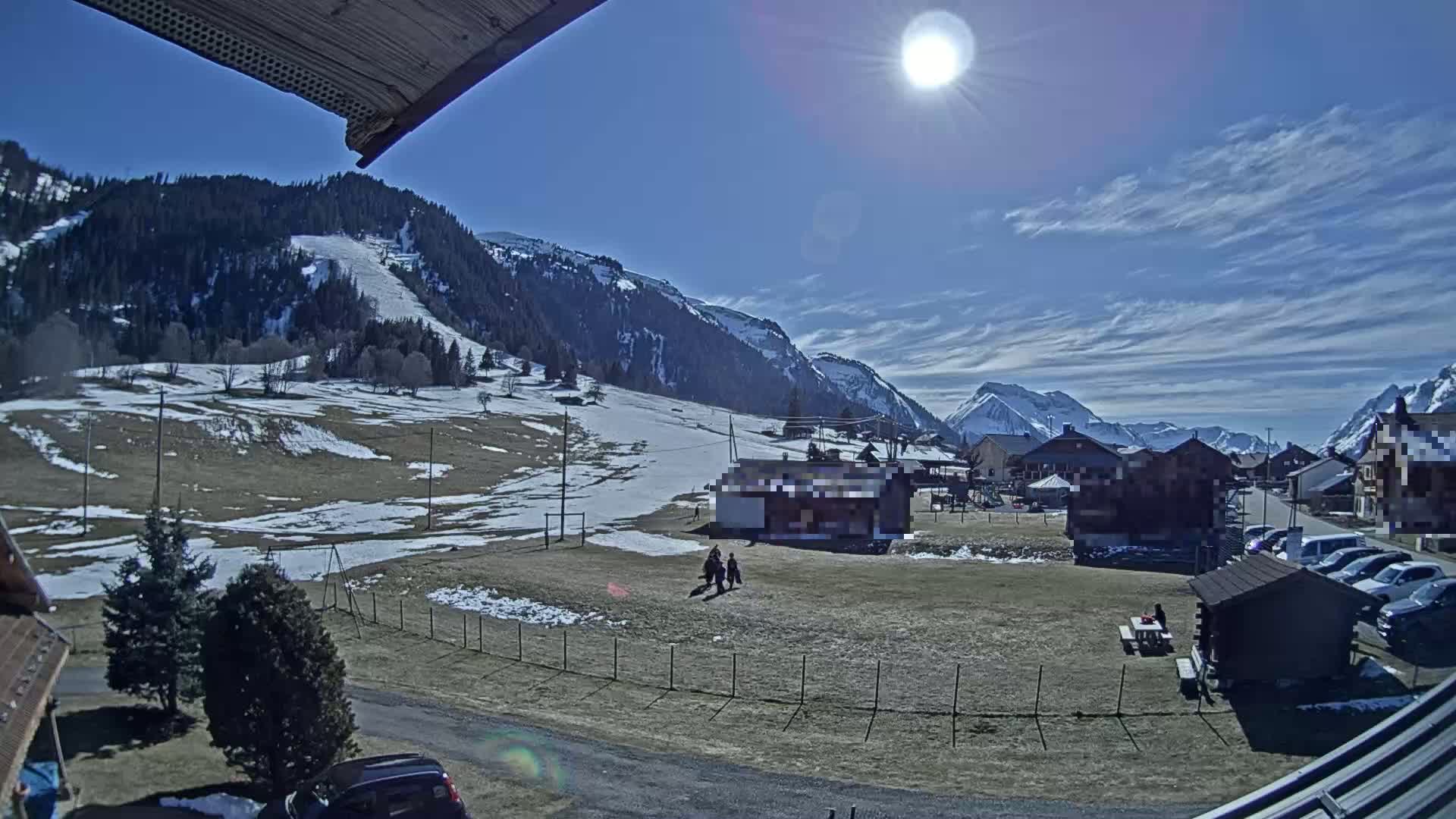 A sunny day in a snow-dusted mountainous region reveals a small village nestled in a valley, with a few people walking near a fenced-in area.