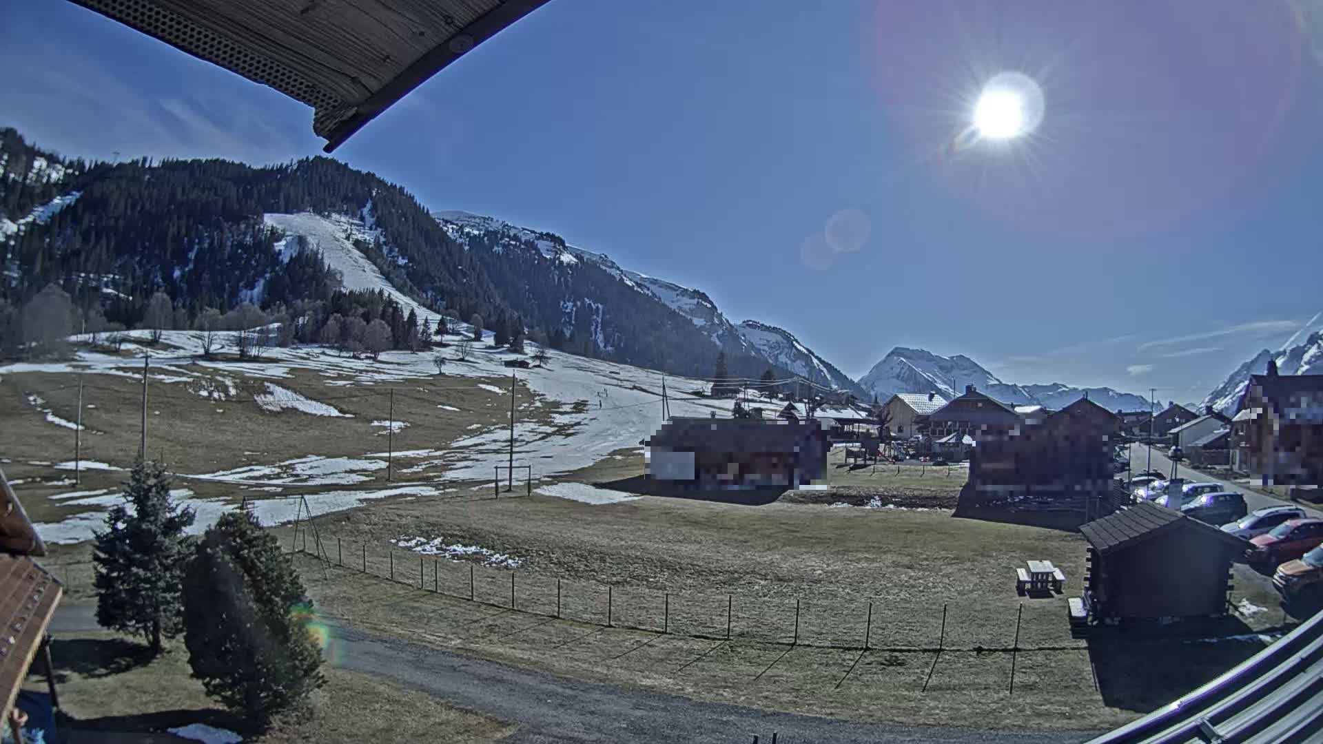 A sunny day in a snow-dusted alpine village nestled among mountains, with patches of snow on the ground and cars parked near some buildings.