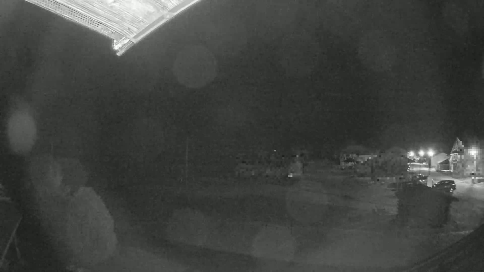 A nighttime, low-light view shows a snow-covered parking lot with buildings and vehicles in the distance under a dark, cloudy sky.