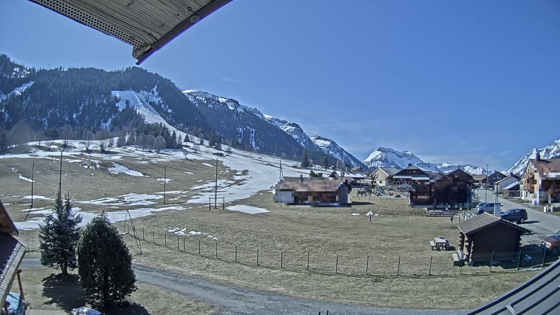 A sunny day reveals a snow-dusted field with a small village nestled beside snow-capped mountains, a ski slope visible on a hillside.