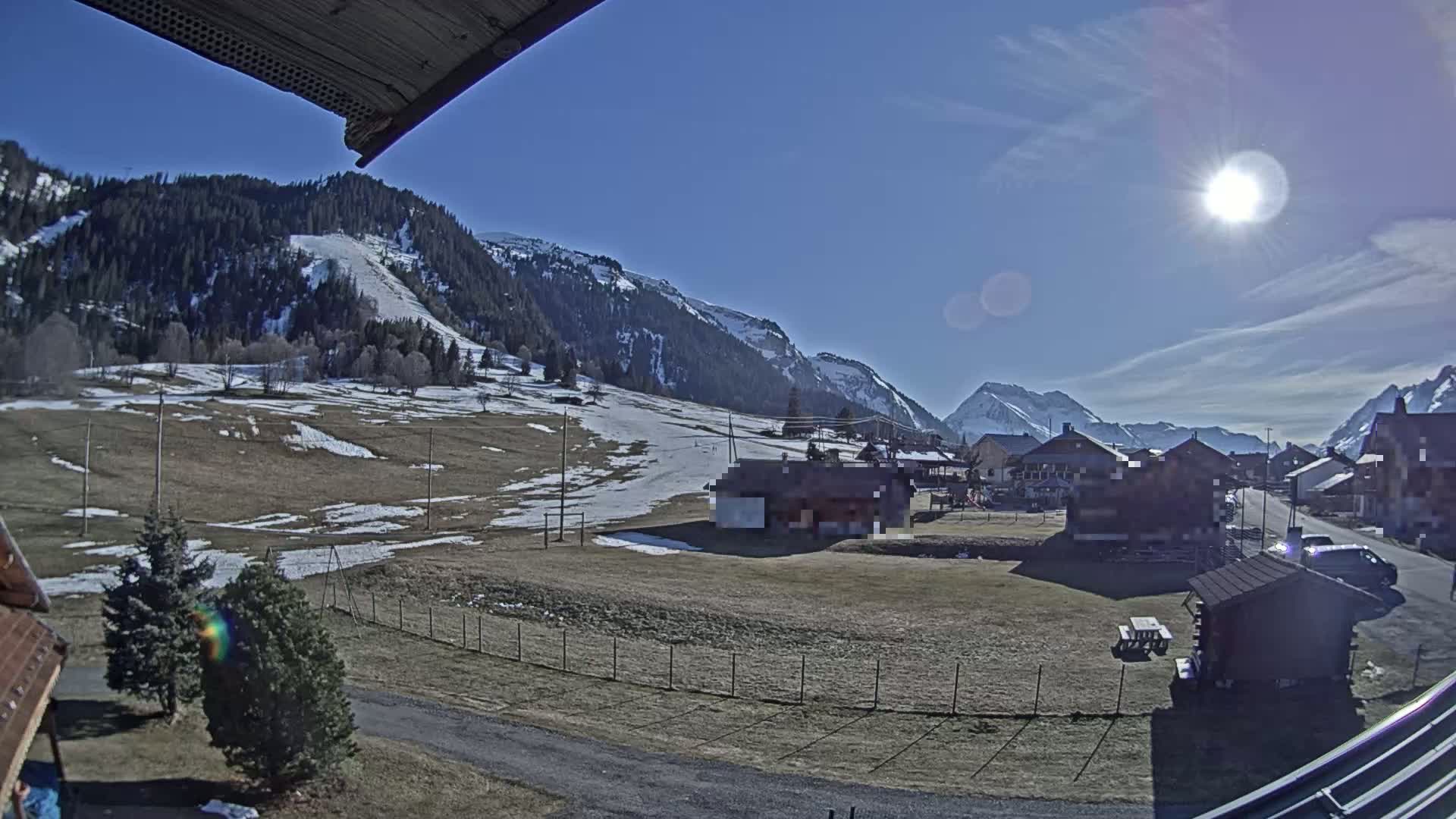 A sunny day in a snow-dusted mountain village shows brown and tan fields, evergreen trees, and several buildings nestled among the foothills.