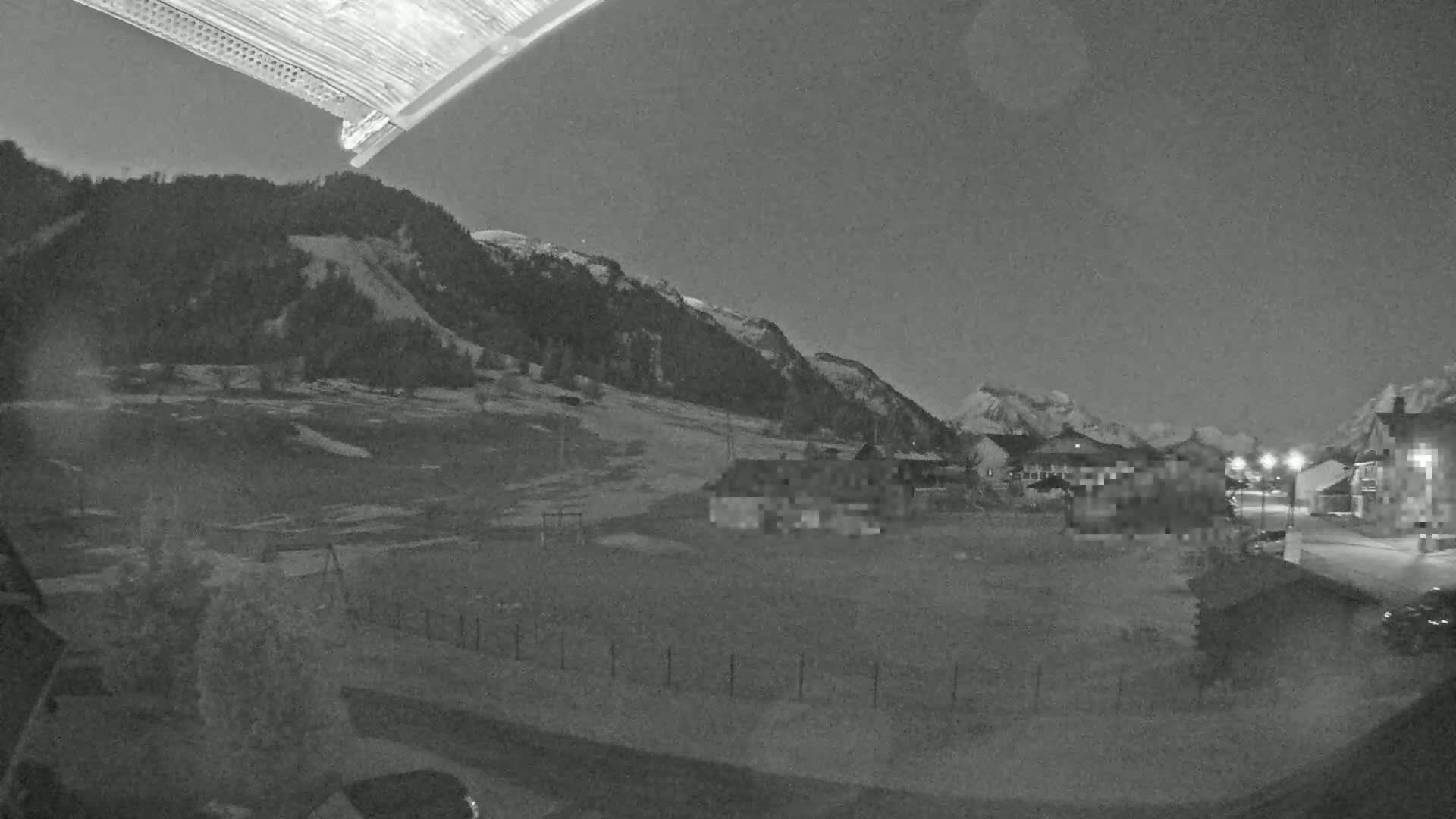 A nighttime view of a snow-covered landscape with a few buildings, a hill, and mountains in the background under a dark sky.