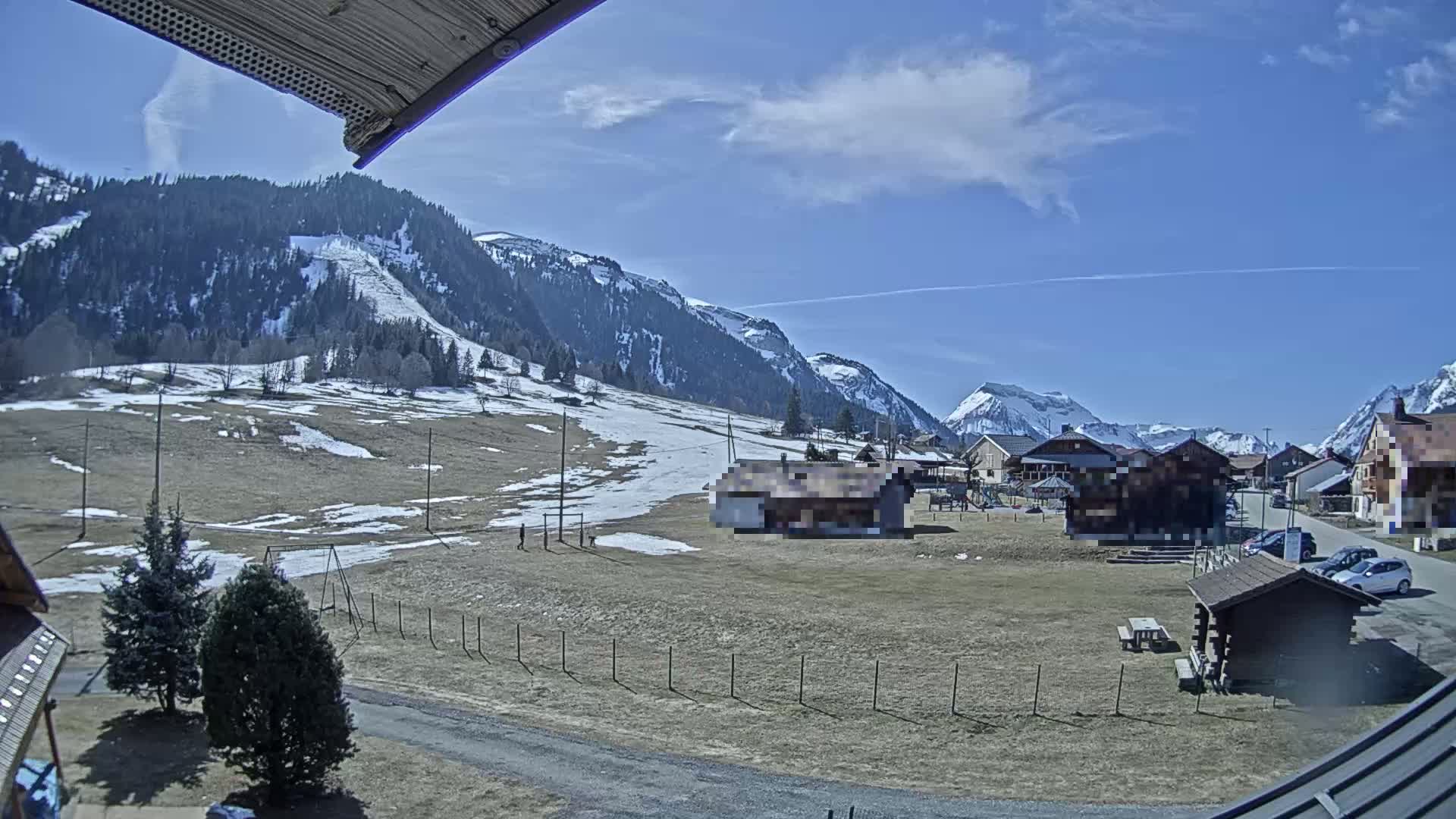A mostly sunny day shows a village nestled in a valley, with snow-dusted mountains in the background and a partially snow-covered field in the foreground.
