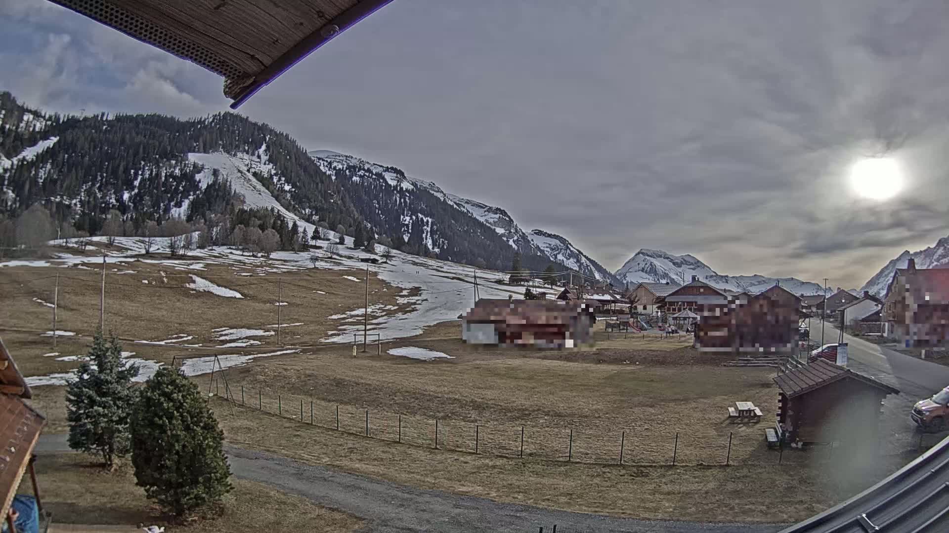 A partly sunny day reveals a mountain village nestled in a valley, with patches of snow visible on the surrounding hills and fields.