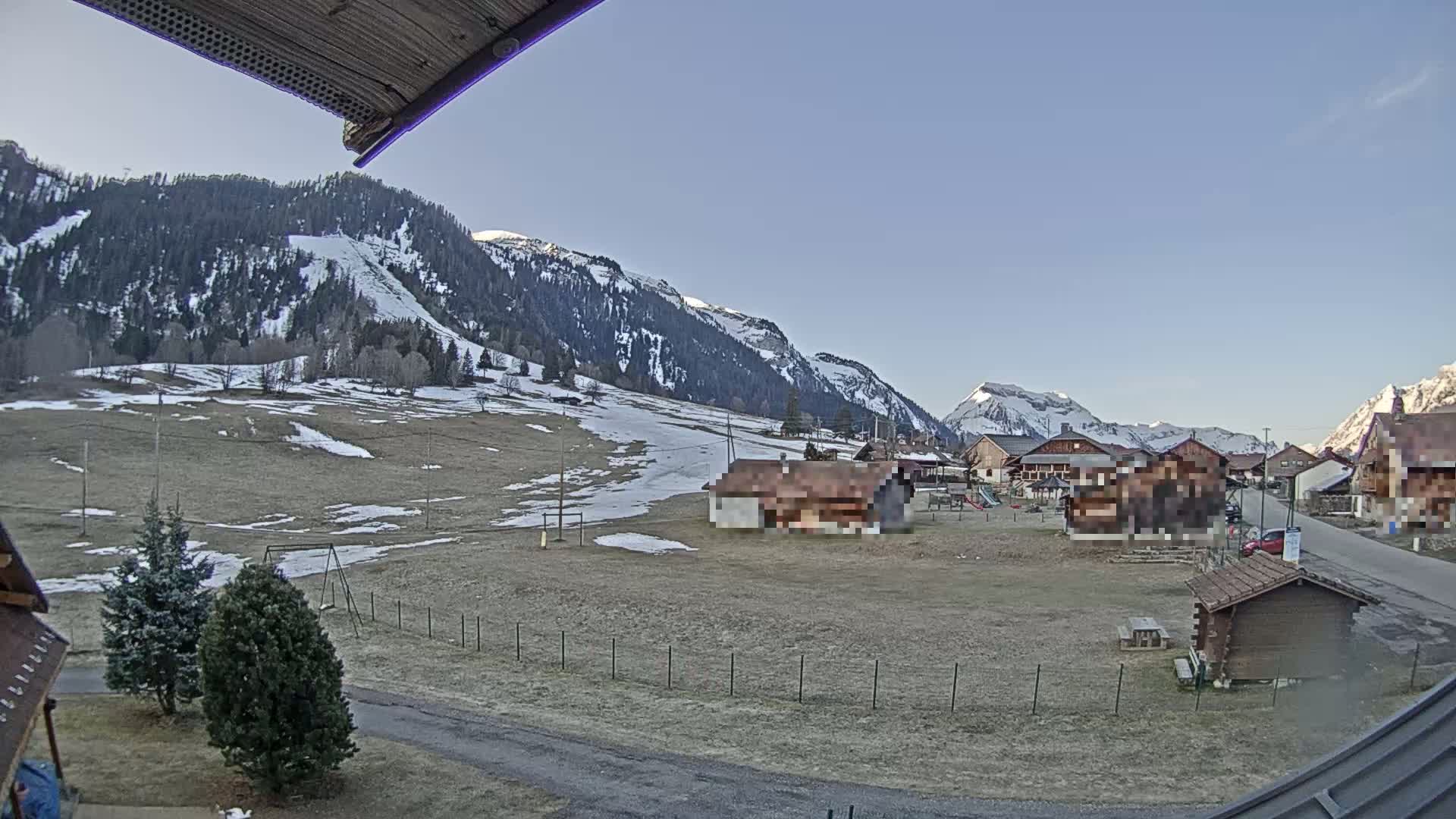 A mostly clear day reveals a small village nestled in a valley, surrounded by snow-dusted mountains and patches of brown and green fields.