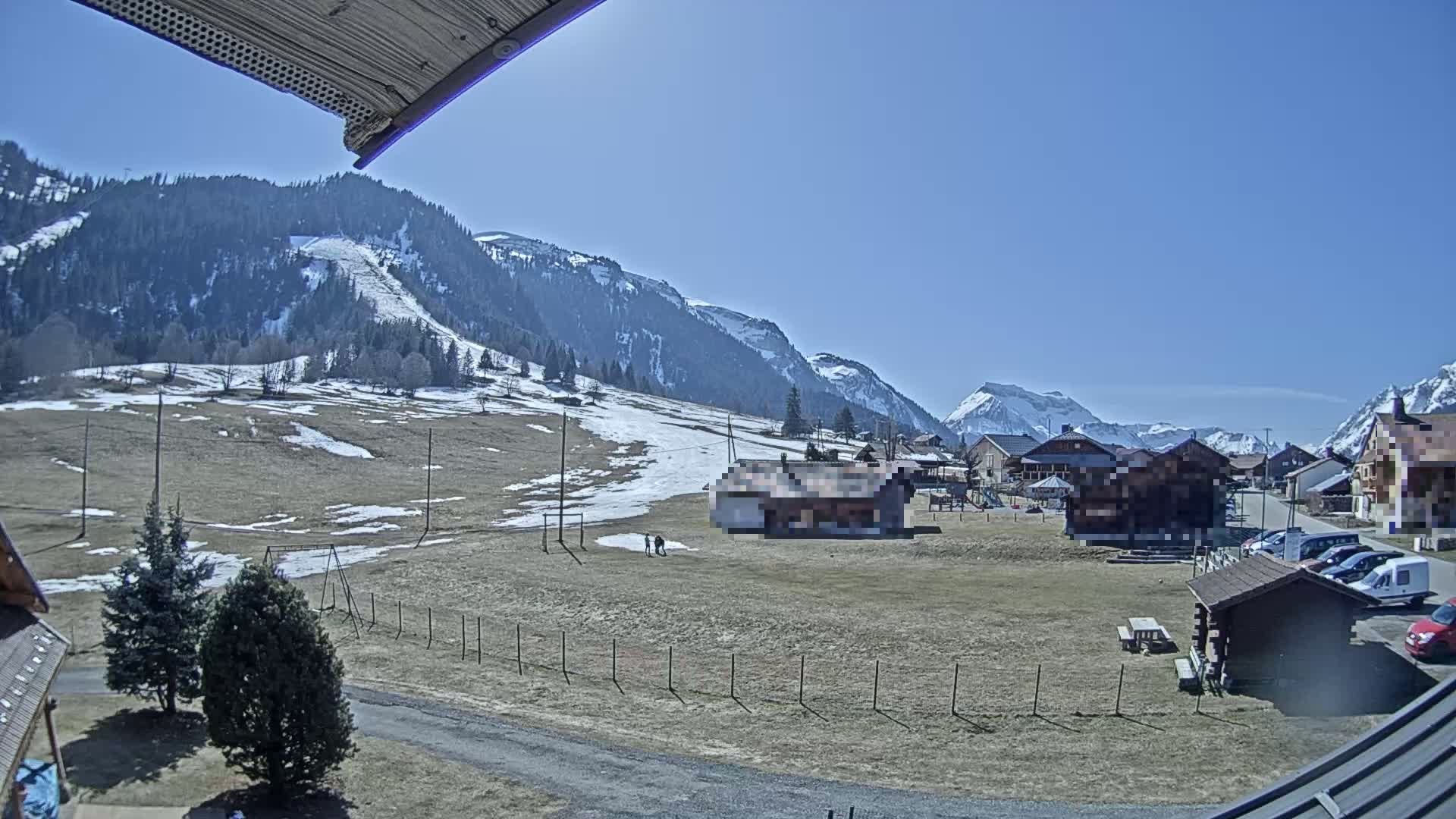 A sunny day reveals a snow-dusted mountainside village nestled amongst the peaks, with a few cars parked near some small buildings and a playground in the foreground.