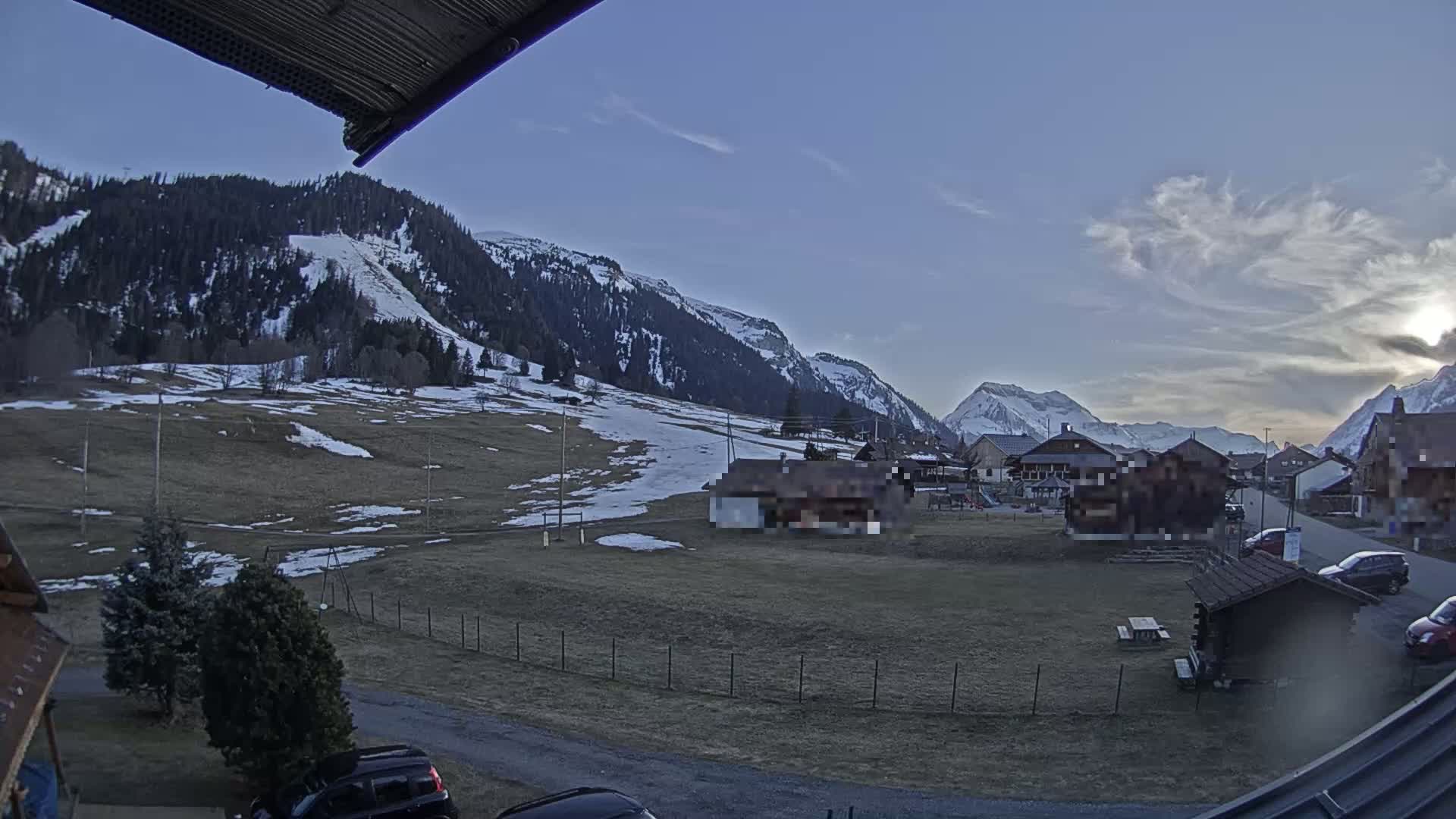 A partly cloudy evening scene shows a snow-dusted grassy field, a small village nestled against snow-capped mountains, and a few cars parked nearby.
