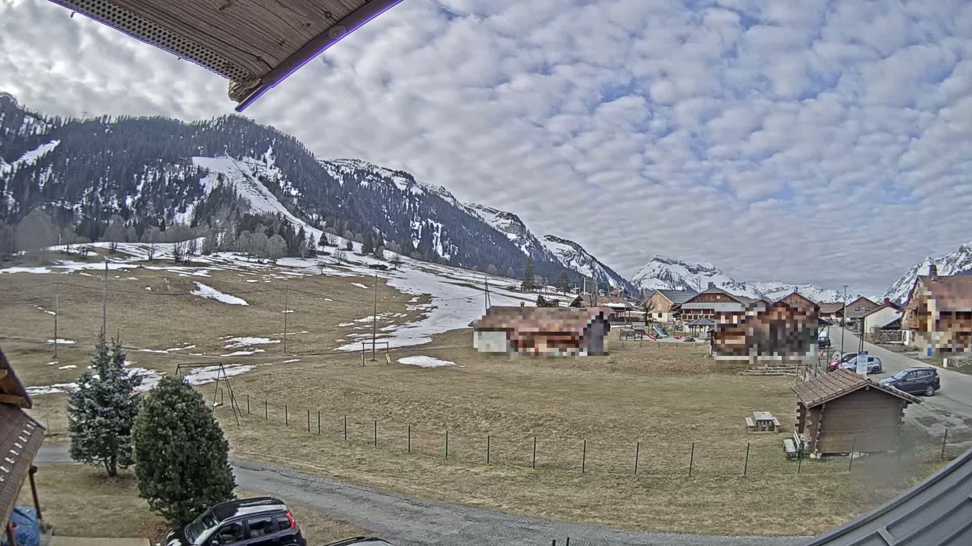 A partly cloudy day reveals a village nestled in a valley between snow-capped mountains, with patches of snow and brown grass visible in the foreground.