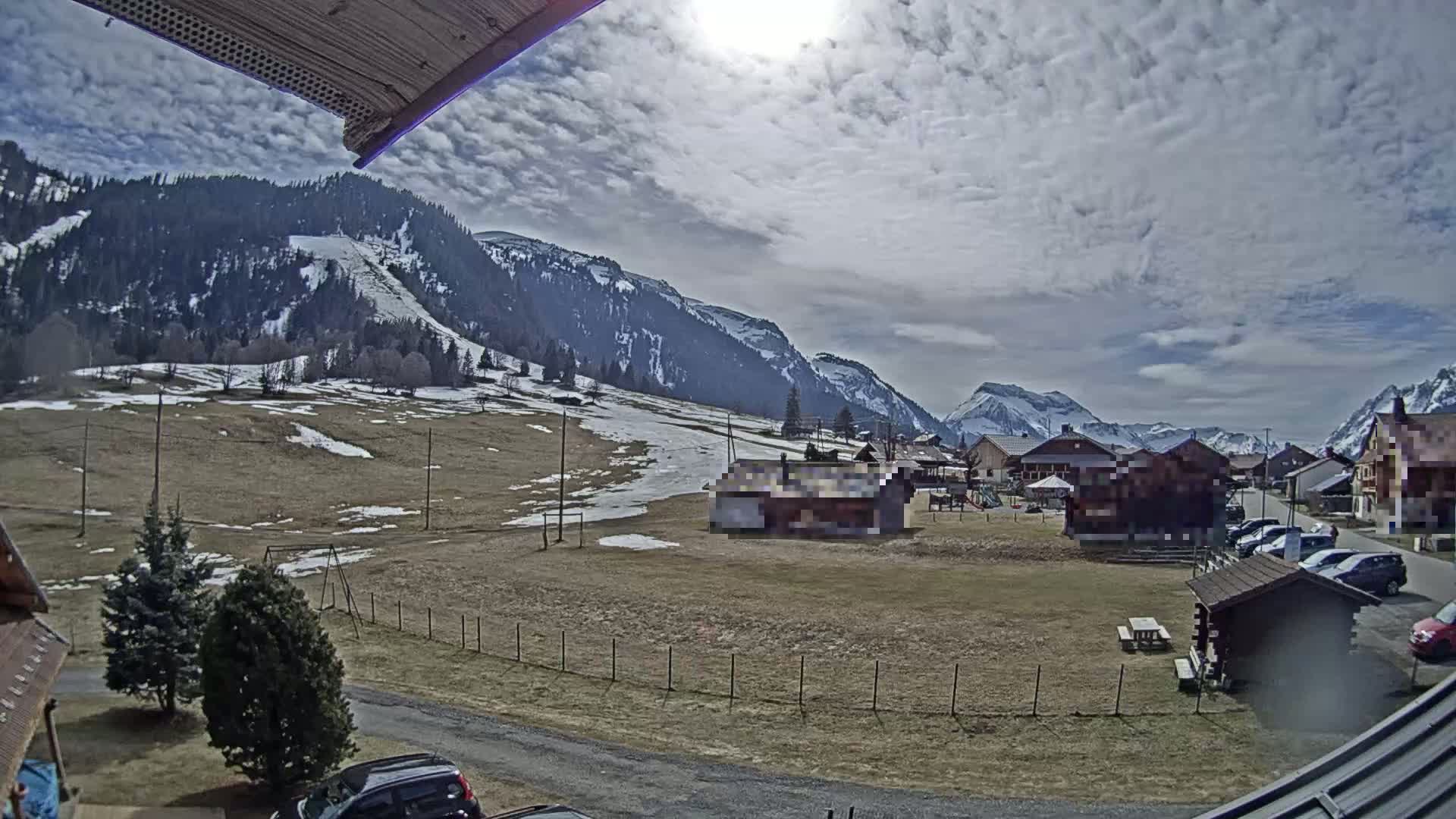 A partially sunny day with cloudy skies shows a mountain village nestled in a valley, with snow-dusted slopes and patches of snow on the ground.