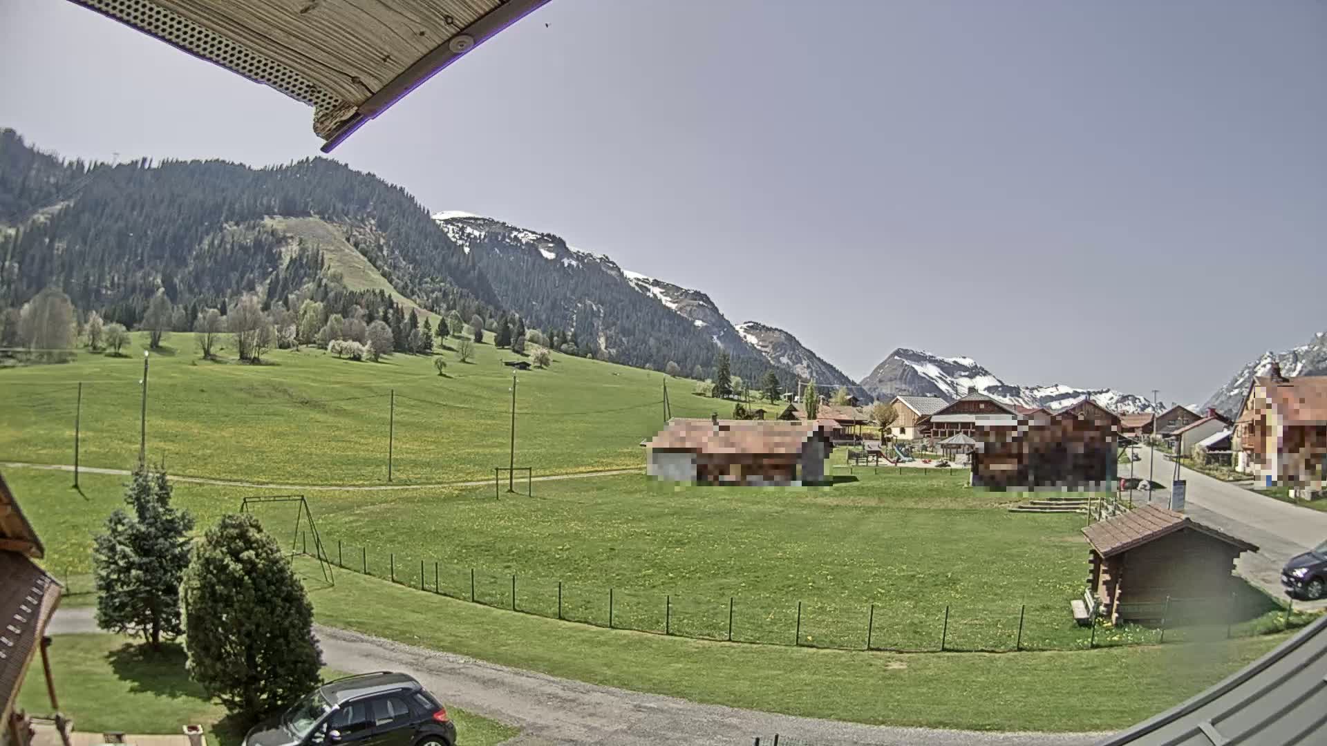 A sunny view of a village nestled in a green valley, surrounded by snow-capped mountains.