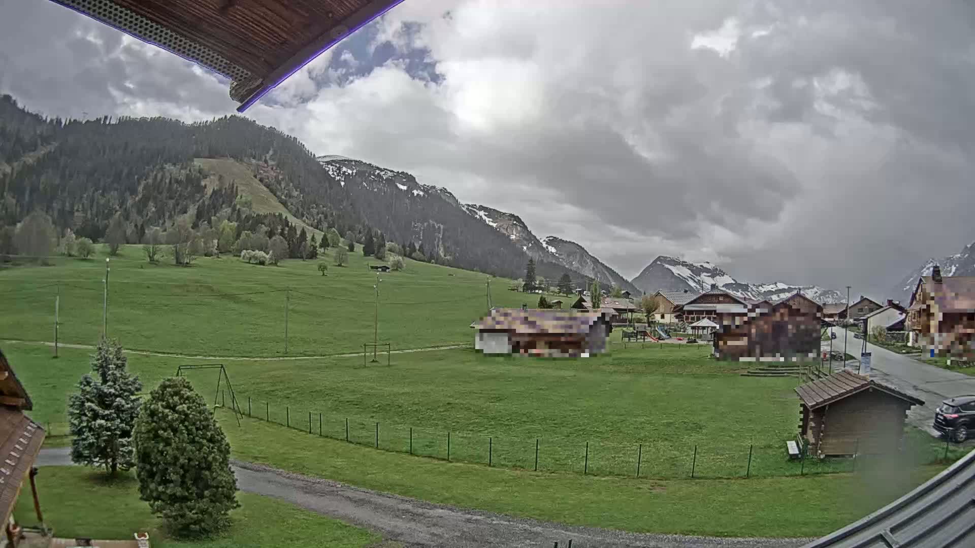 A grassy field with a few buildings and a swing set sits before snow-capped mountains under an overcast sky.