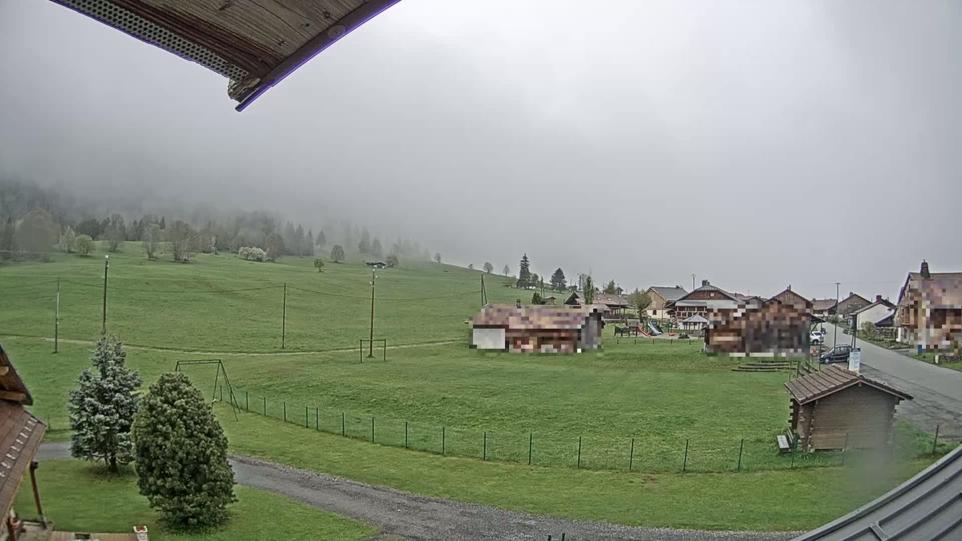 A foggy, overcast day reveals a grassy field with a few buildings, trees, and a playground, all nestled in a valley surrounded by a forest.