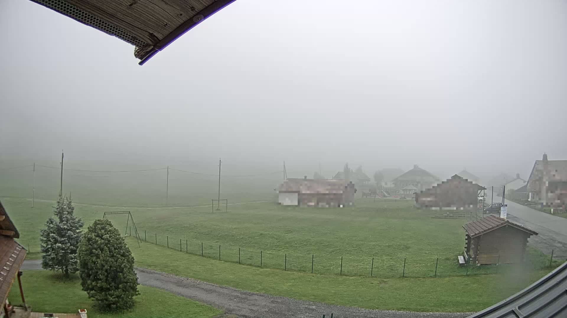 A foggy view of a grassy field with several buildings and a swing set, seen from a slightly elevated vantage point.