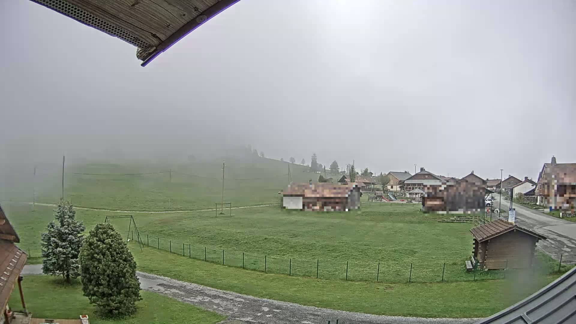 A foggy, green field with a few buildings and a playground in the background, viewed from a slightly elevated angle.