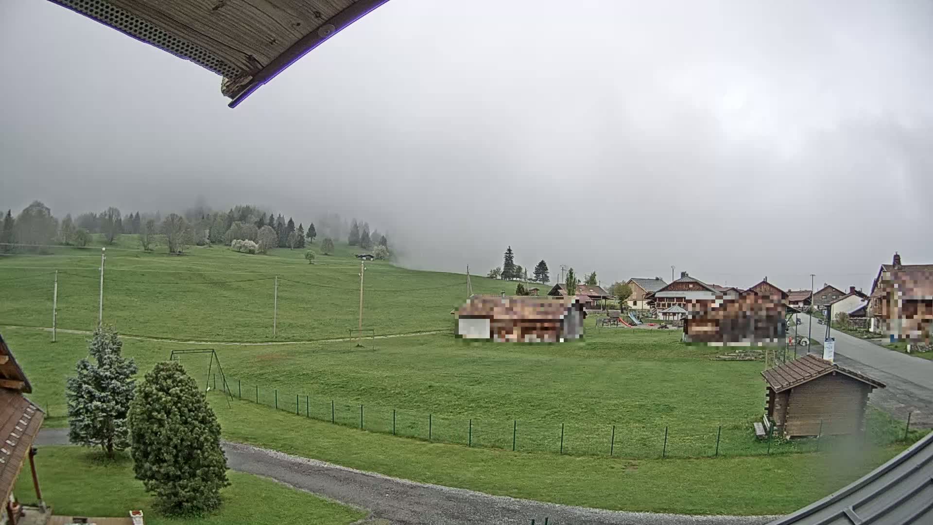 A foggy, overcast day reveals a grassy hillside with a small village in the distance, featuring several wooden buildings and a playground.