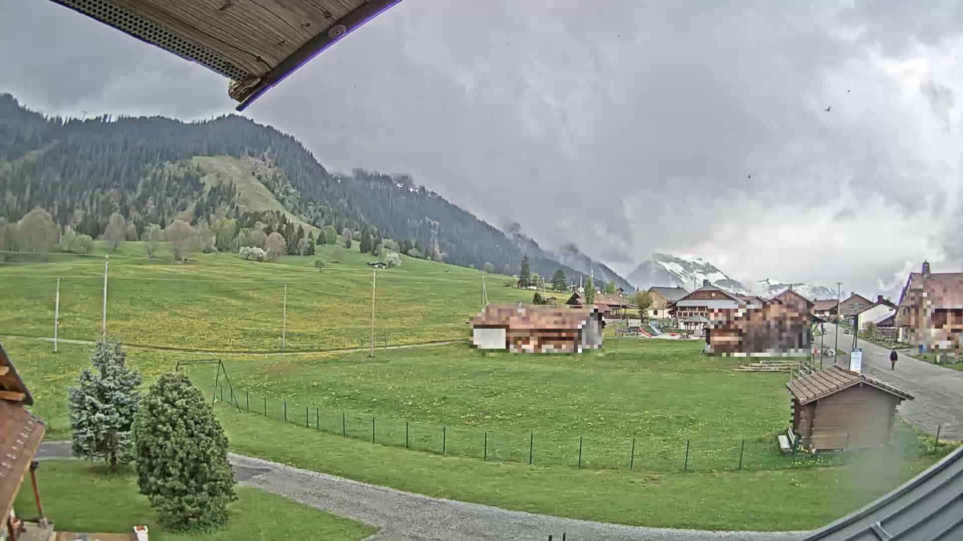 A view of a green valley with a few buildings and trees under an overcast sky, backed by mountains partially covered in snow.