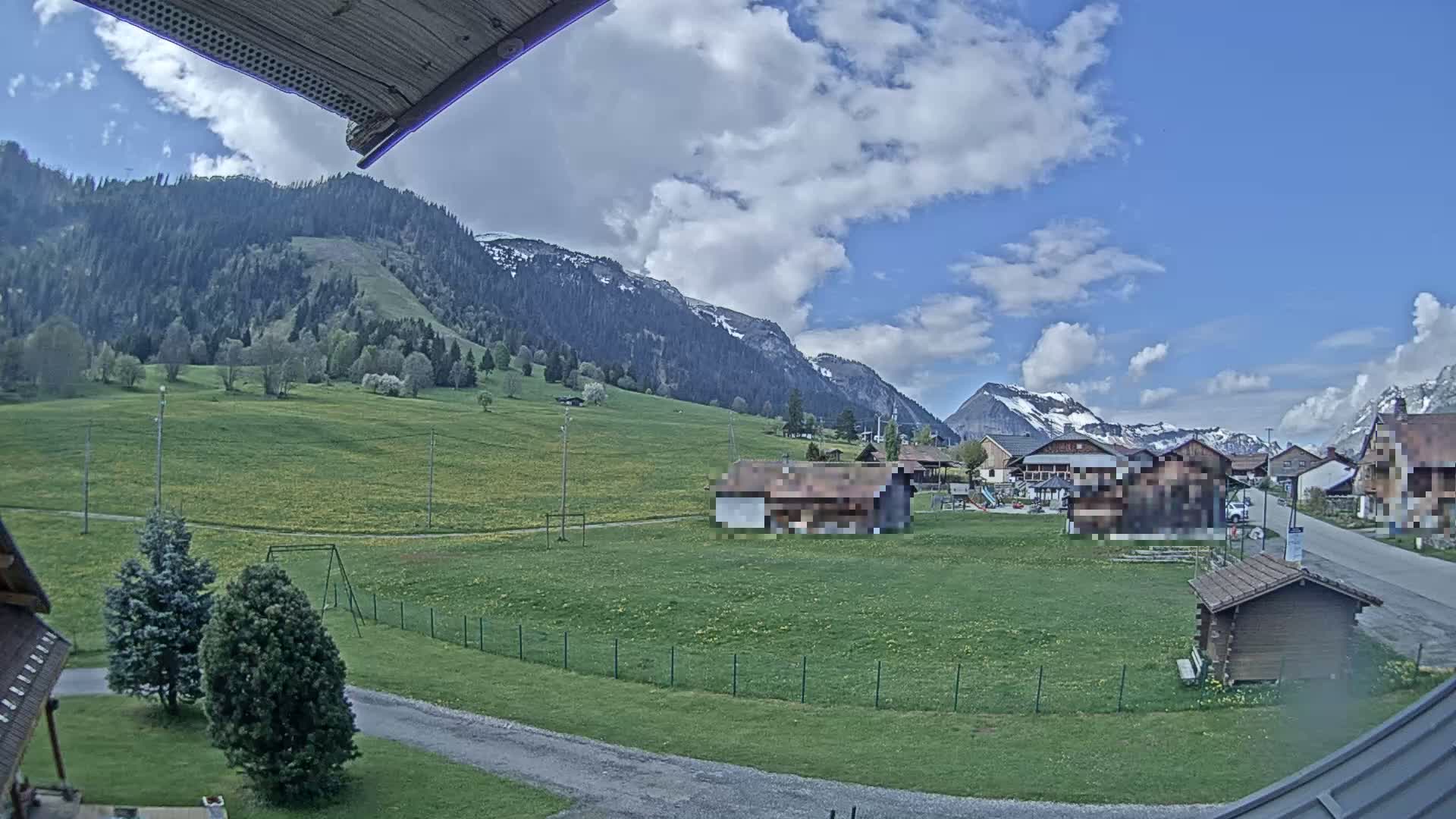 A partly cloudy day reveals a village nestled in a green valley, surrounded by mountains with some snow visible on the peaks.