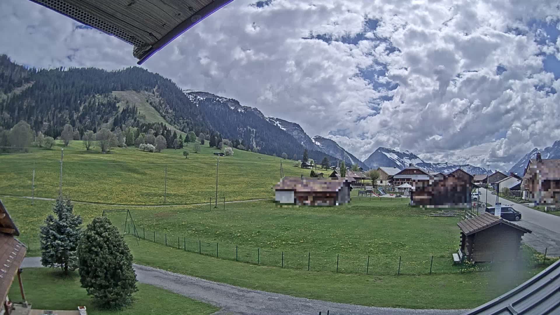 A partly cloudy day reveals a mountainous landscape with a village nestled in a green valley, featuring a few buildings and grassy areas.