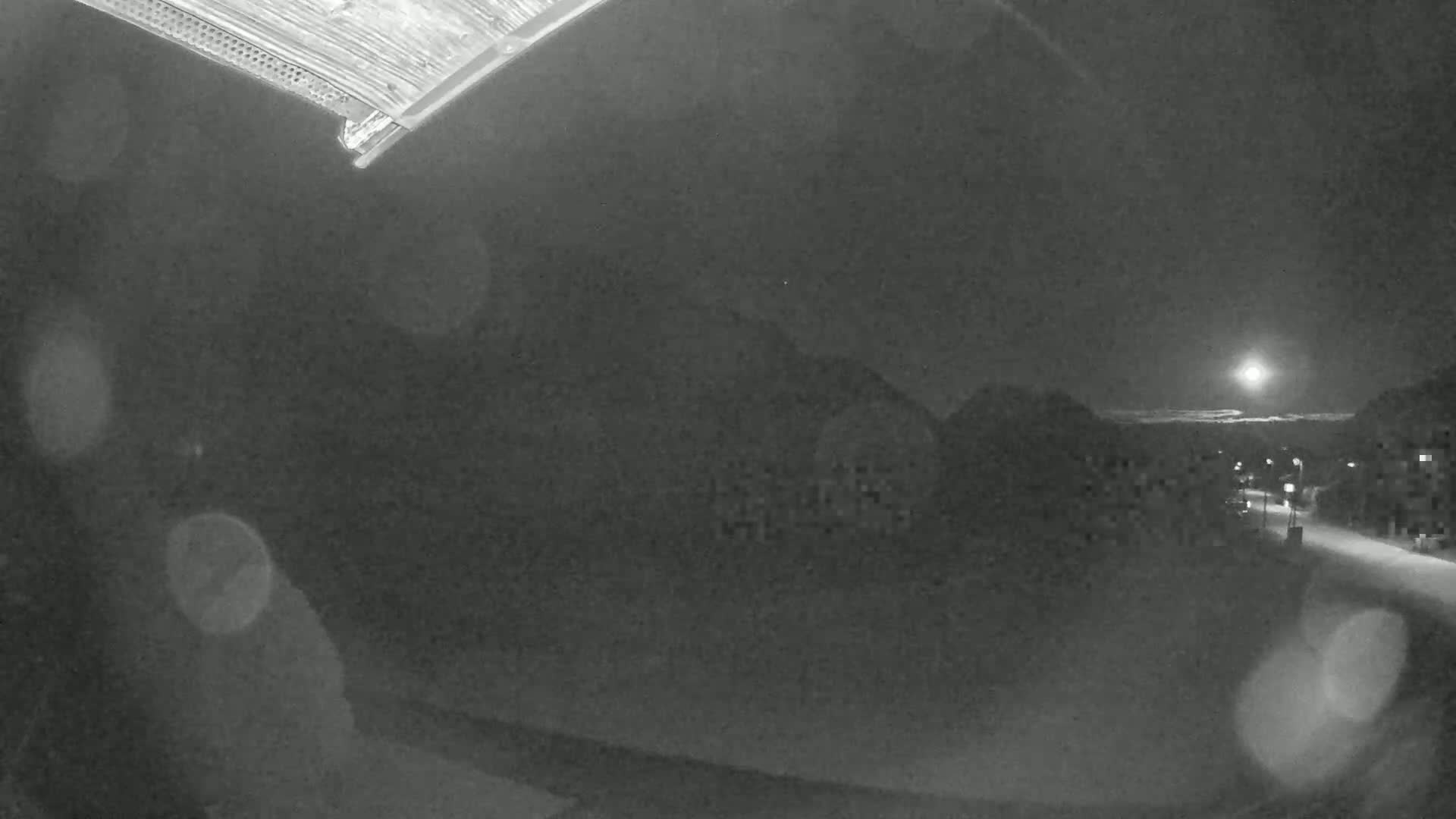A nighttime, snowy scene shows a moonlit street with houses and hills in the distance, viewed from what appears to be a window.