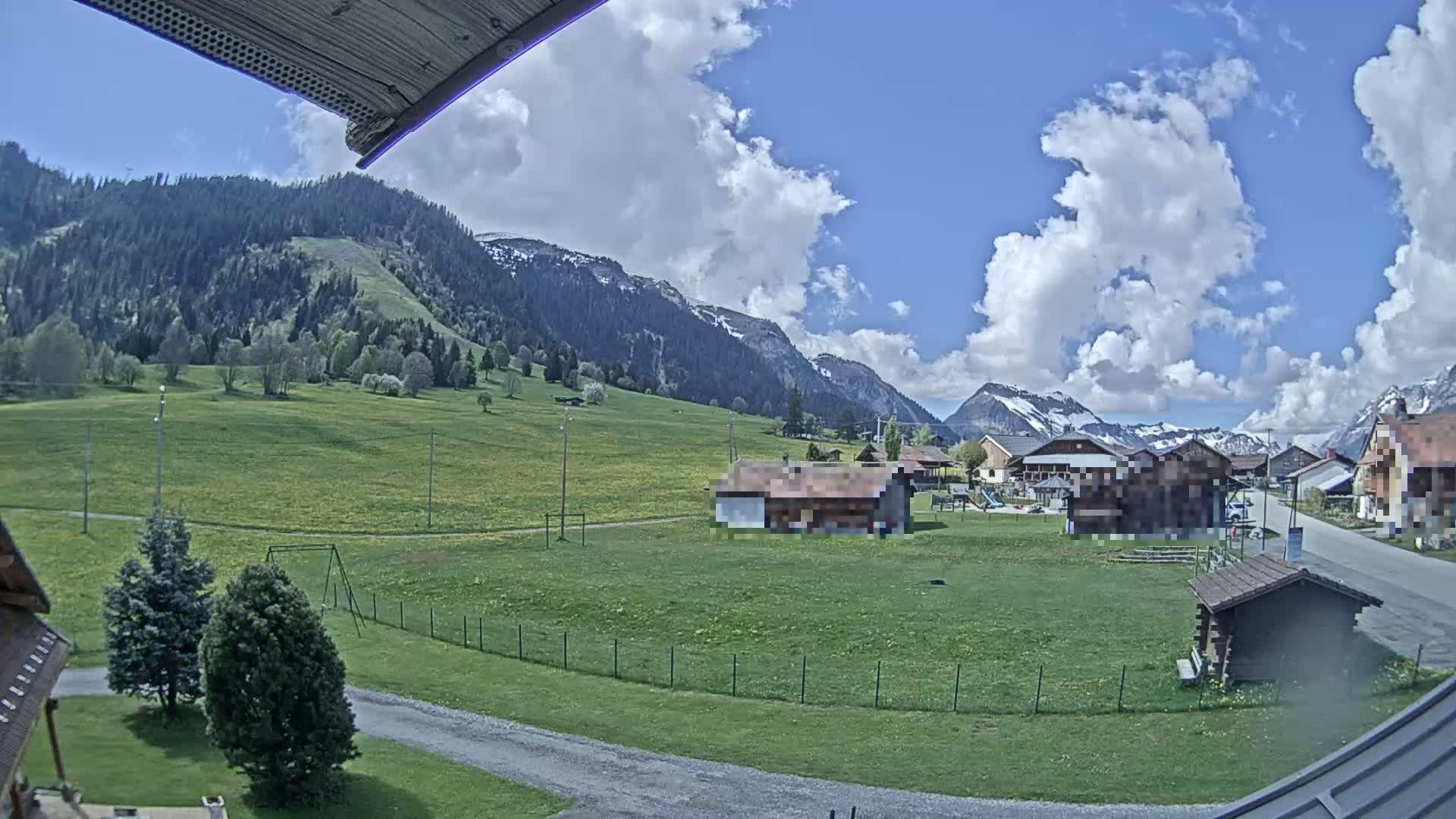 A partly cloudy day reveals a mountainous landscape with a small village nestled in a grassy valley, featuring several buildings and a playground.