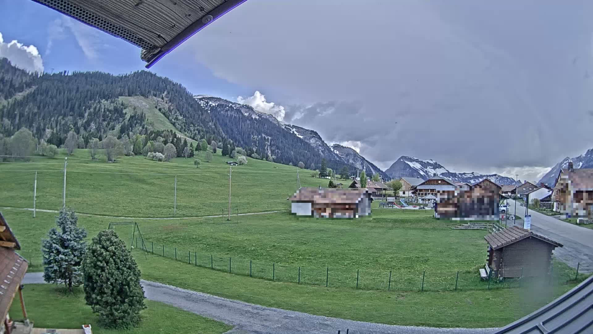 A grassy valley with a few buildings and a playground is nestled between snow-capped mountains under a mostly cloudy sky.