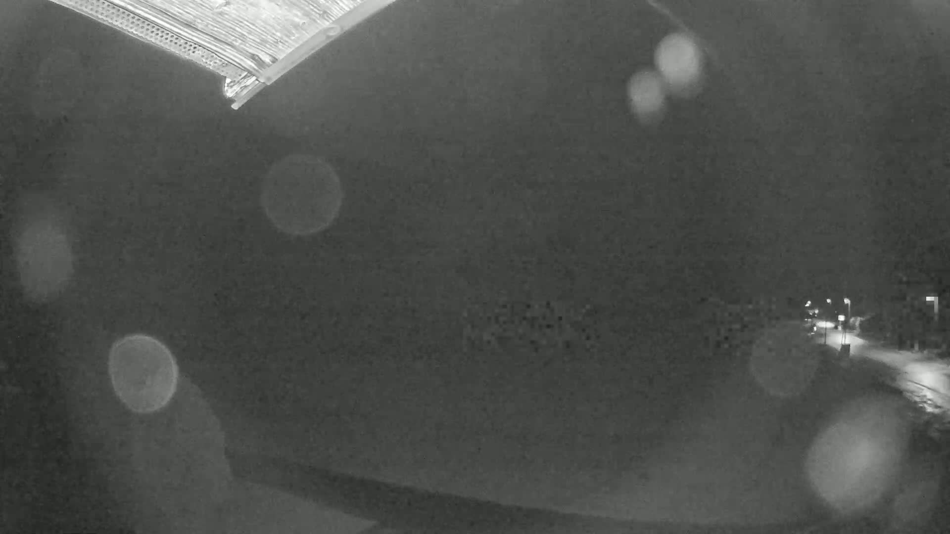 A blurry nighttime view of a snow-covered road with dimly lit buildings in the distance, seen from a high angle with rain or snow on the camera lens.