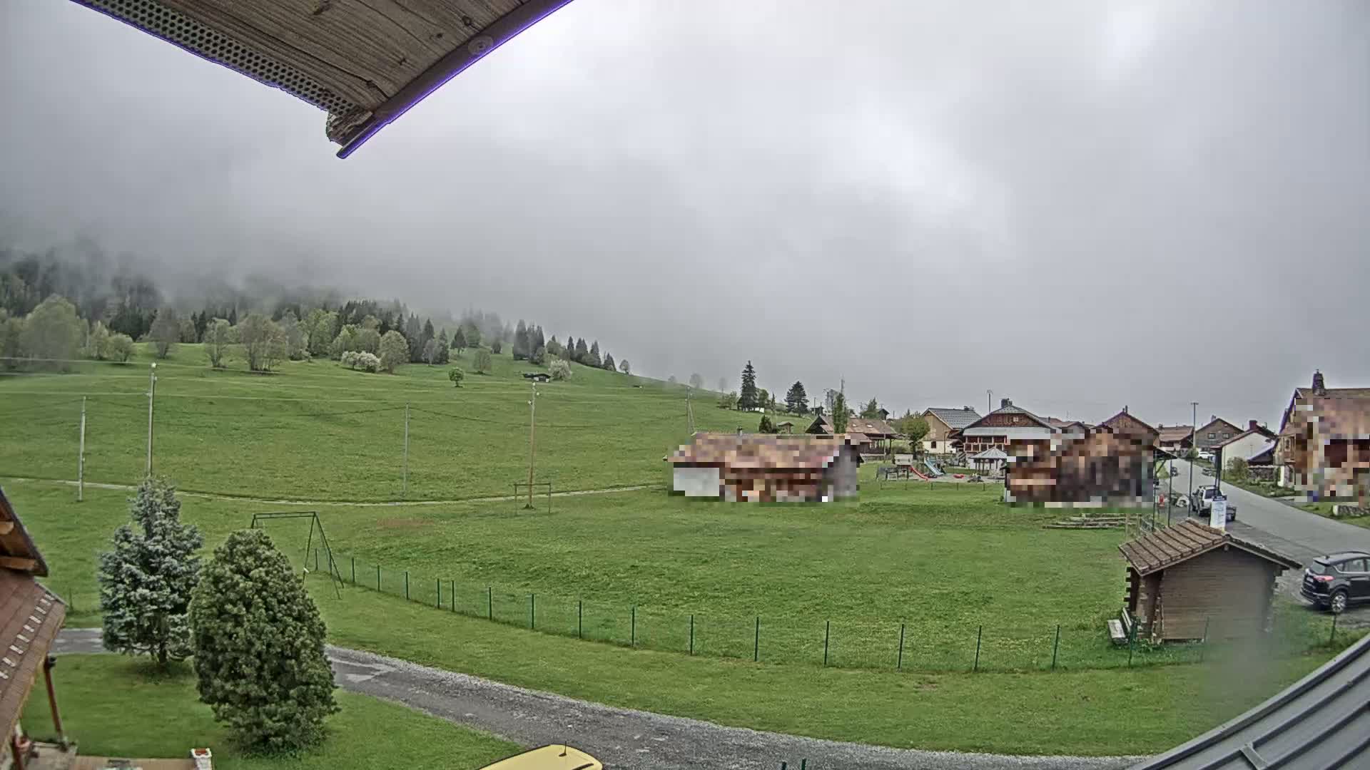 A misty, overcast day reveals a grassy expanse, a small village of houses, and a playground, nestled among forested hills.