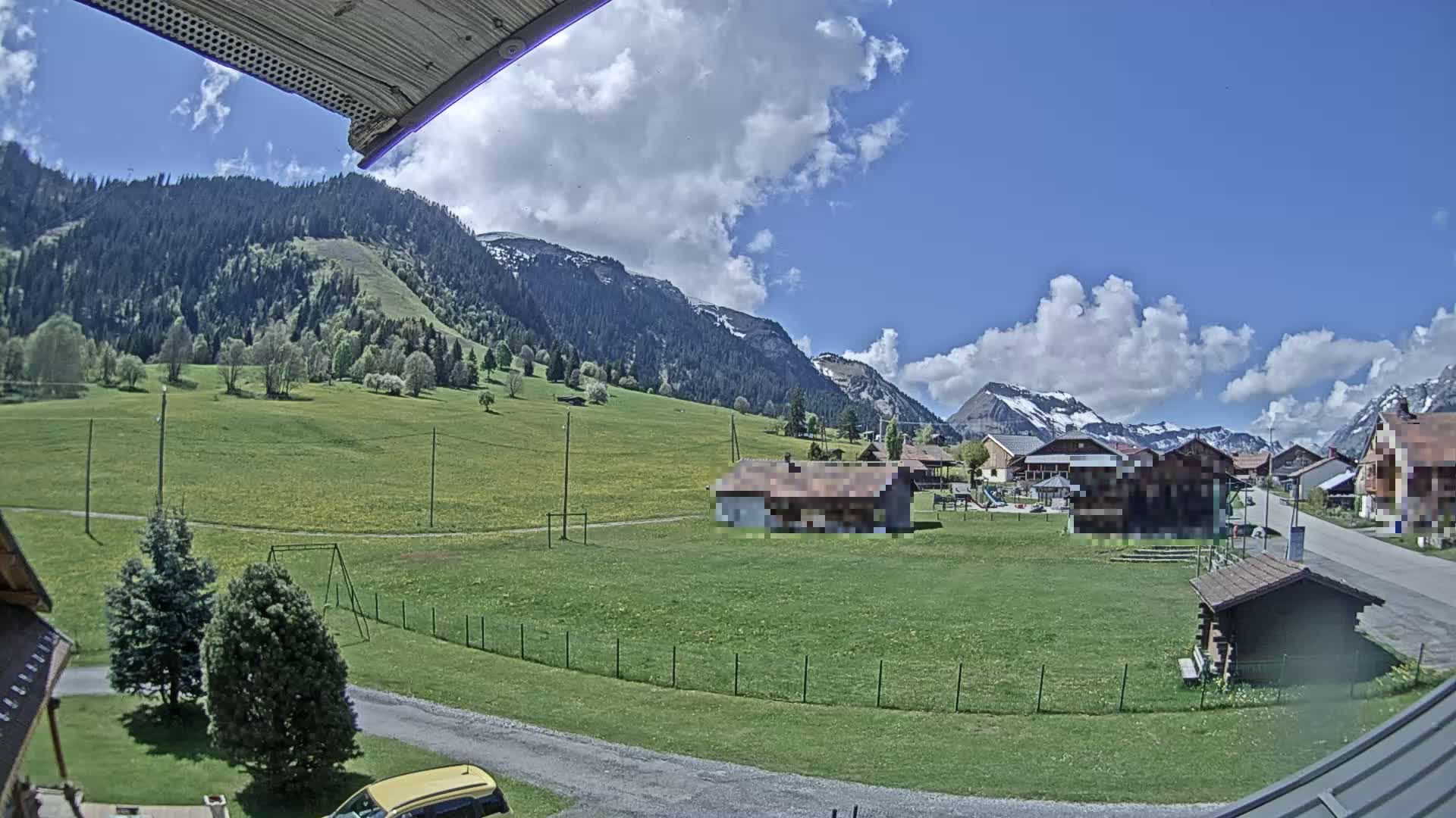 A partly cloudy, sunny day reveals a village nestled in a green valley surrounded by mountains, some with snow-capped peaks.