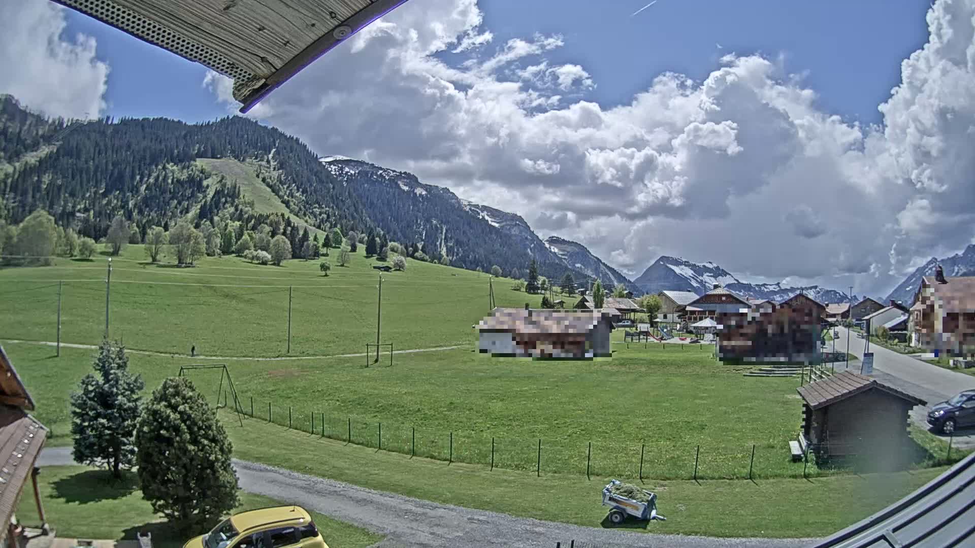 A partly cloudy day reveals a green valley nestled between mountains, with a small village and playground visible in the distance.