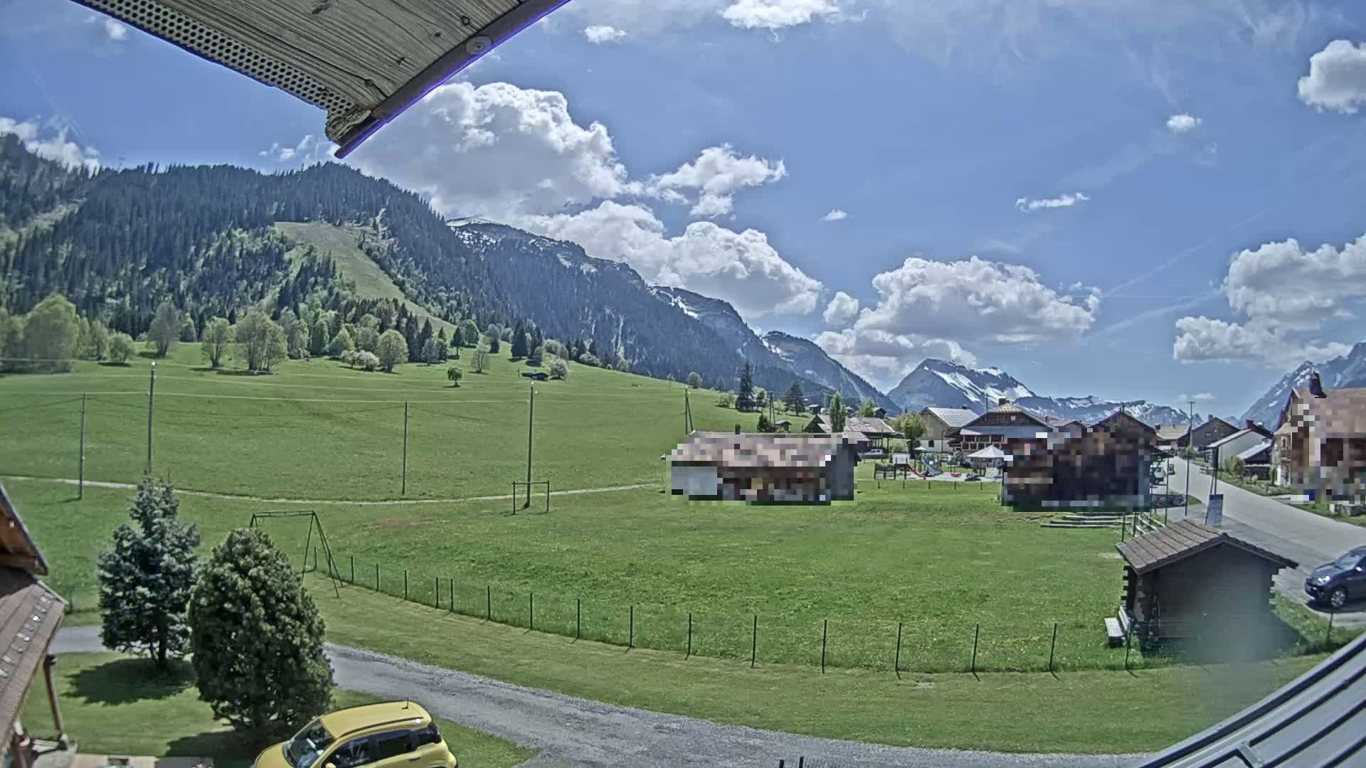 A partly cloudy day reveals a small village nestled in a green valley, with mountains in the background.