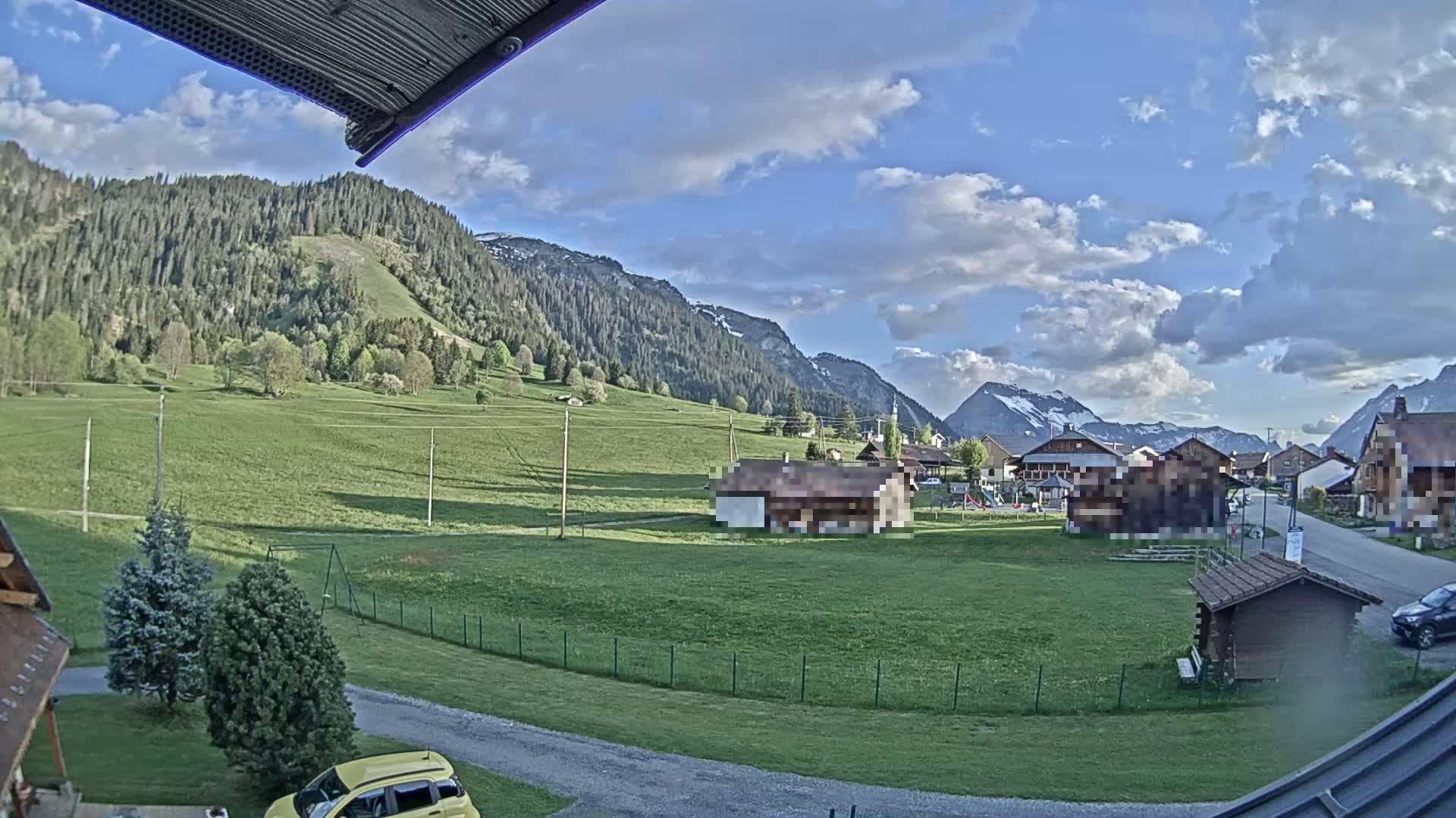 A partly cloudy afternoon view of a village nestled in a valley between green hills and snow-capped mountains.