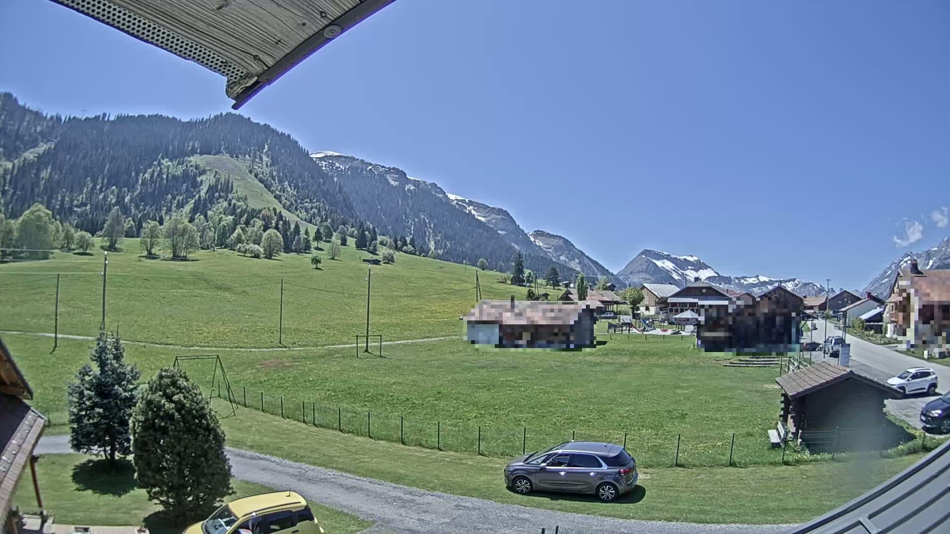 A sunny day reveals a village nestled in a green valley, surrounded by mountains with patches of snow, and cars parked near a grassy area with a swing set.