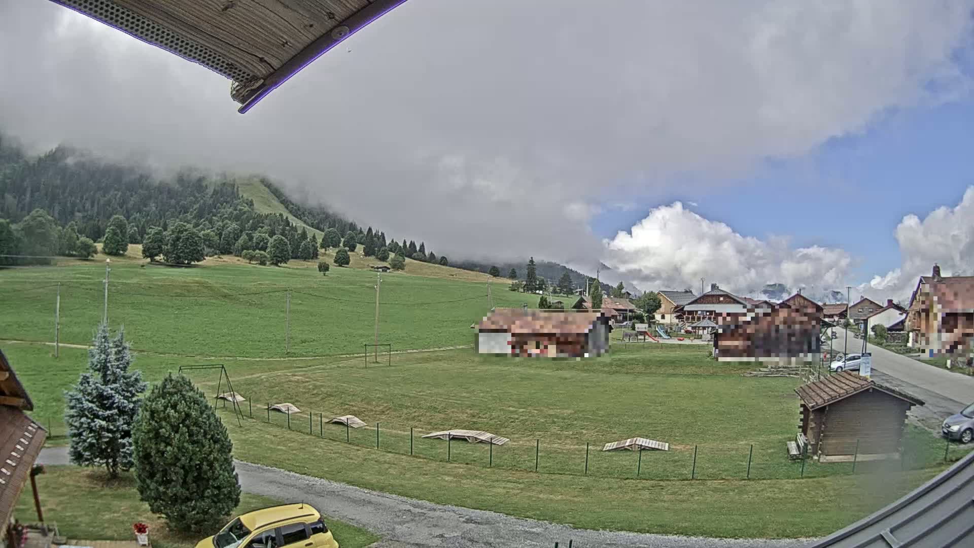A partly cloudy day reveals a village nestled in a valley, with green fields, a few buildings, and a mountain shrouded in mist in the background.