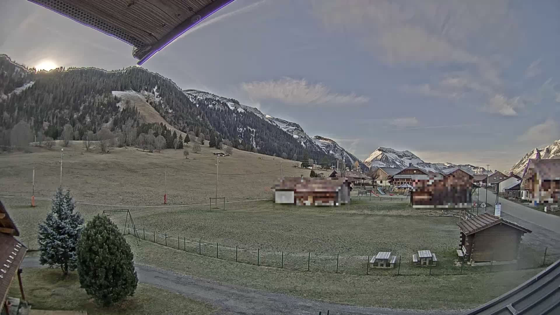 A wide-angle landscape captures a frosty green valley with a village and scattered trees, set against snow-capped mountains under a partially cloudy sky with the sun visible on the left.