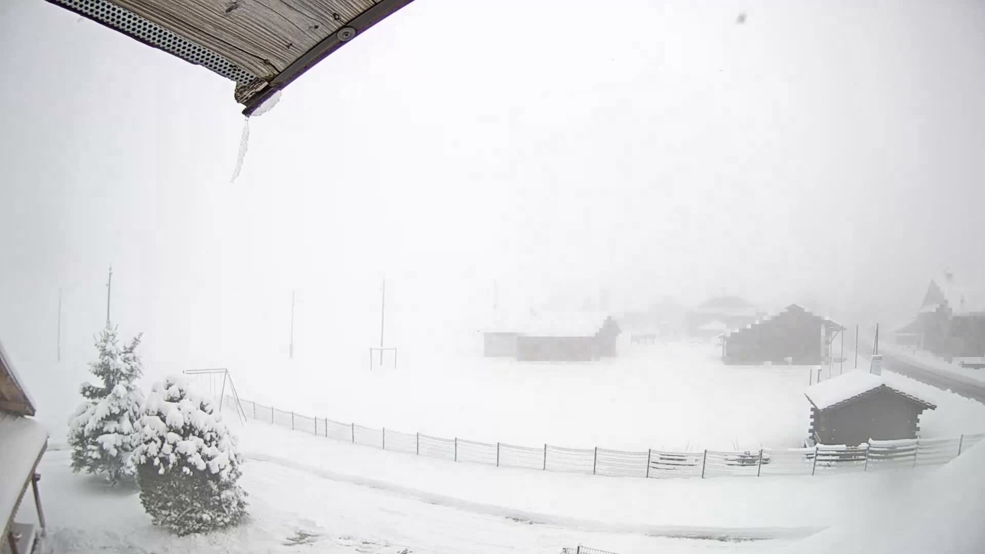 A severe winter whiteout covers a snowy landscape featuring obscured buildings, snow-laden trees, and an icicle dangling from a wooden eave in the foreground.