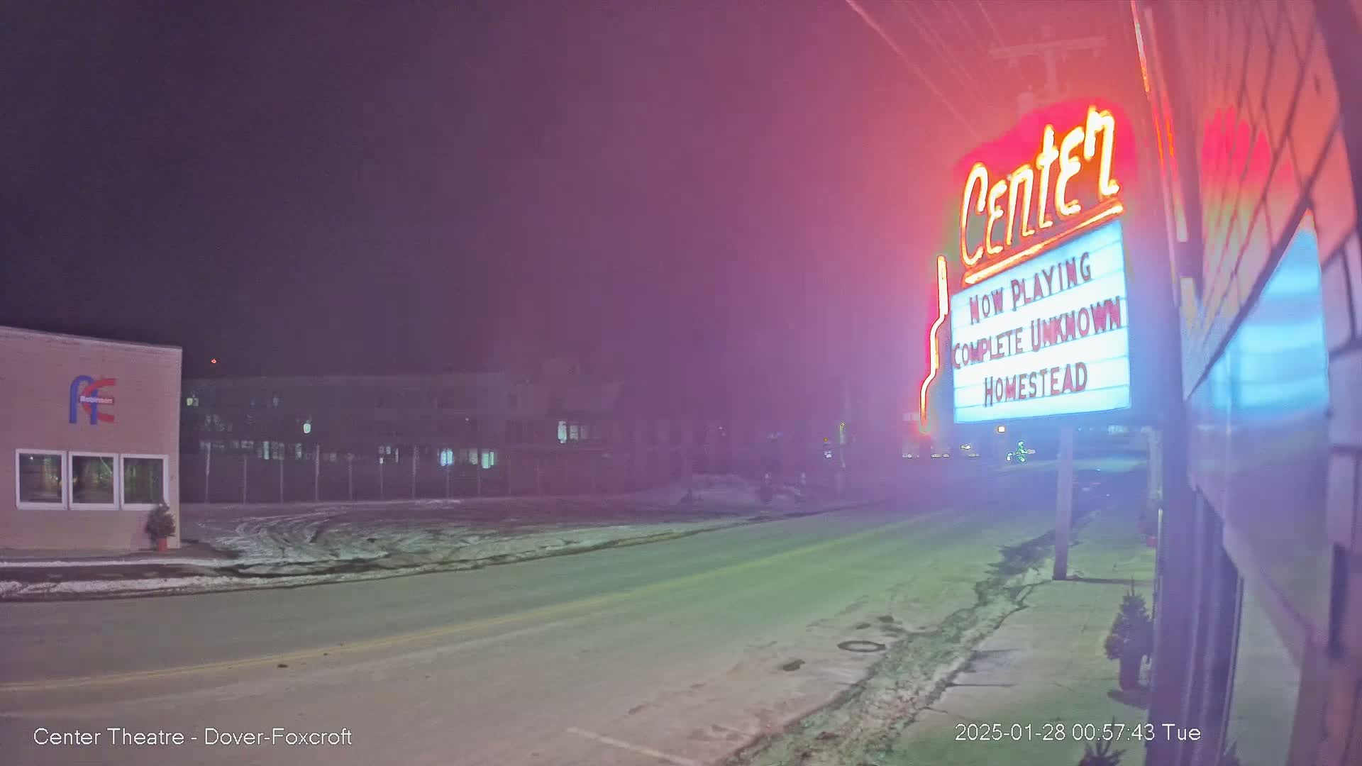 A nighttime street scene with light snow on the ground, showing a movie theater marquee advertising "Complete Unknown" and "Homestead," under hazy, pinkish-red lighting.