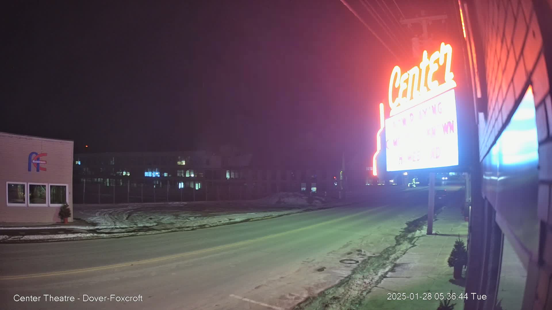A nighttime street scene with snow on the ground shows a neon sign for a theater, a building with windows, and other buildings in the distance under a dark sky.